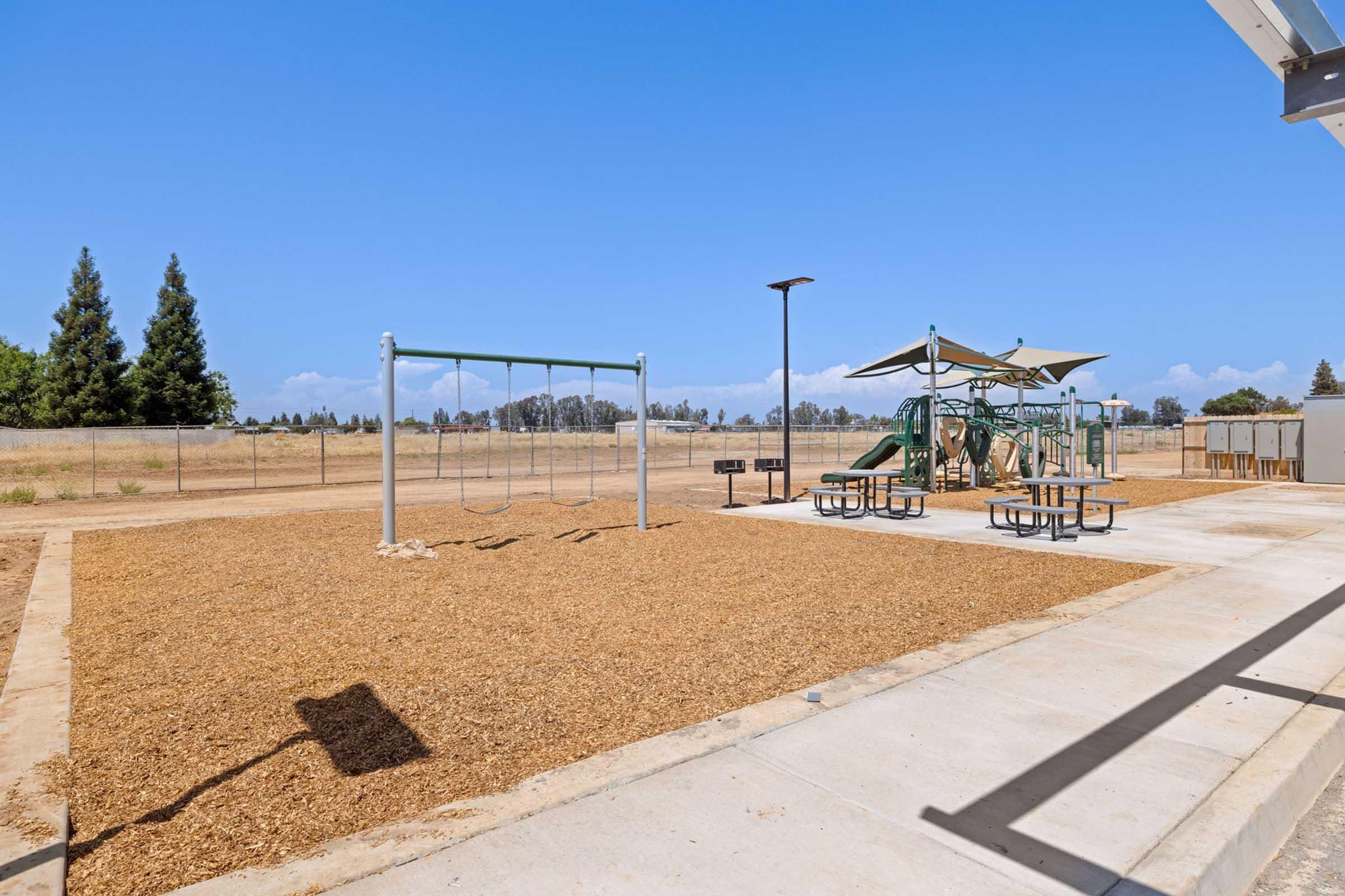 A playground area featuring swings and a slide, surrounded by mulch. Picnic tables are positioned nearby under shade structures. The background includes trees and an open field, with clear blue skies overhead.