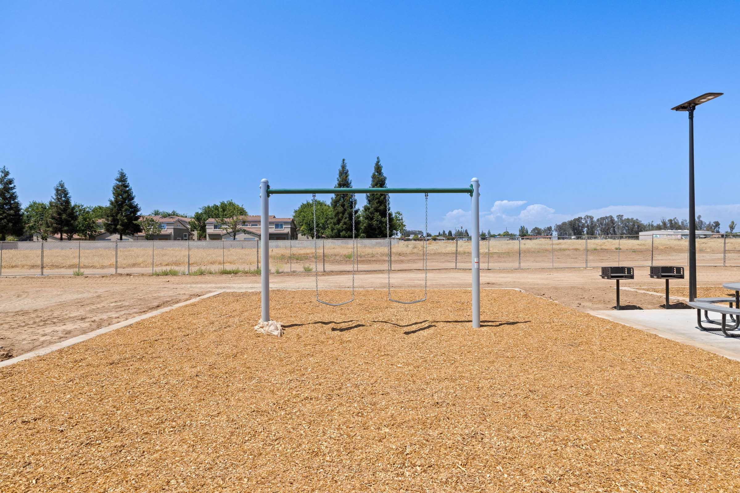 A swingset in a playground, surrounded by wood chip mulch. In the background, green trees and a fenced area are visible under a clear blue sky, with a few picnic tables and grills nearby. The playground area appears empty, indicating a peaceful, open space.