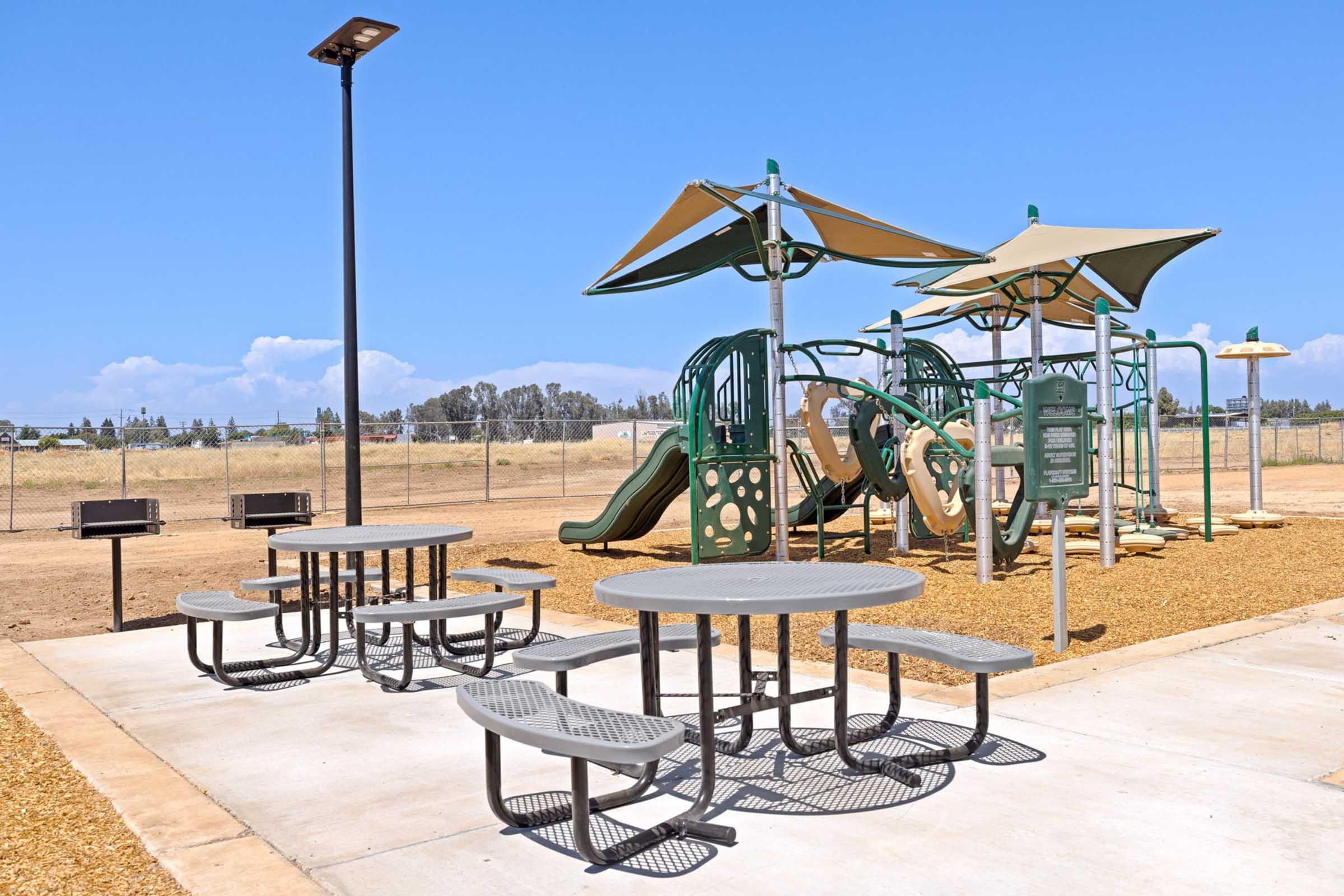 A playground area featuring green slides, climbing structures, and shaded seating tables on a sunny day. The environment is open with a clear blue sky and a fence in the background, indicating a recreational space designed for children and families.