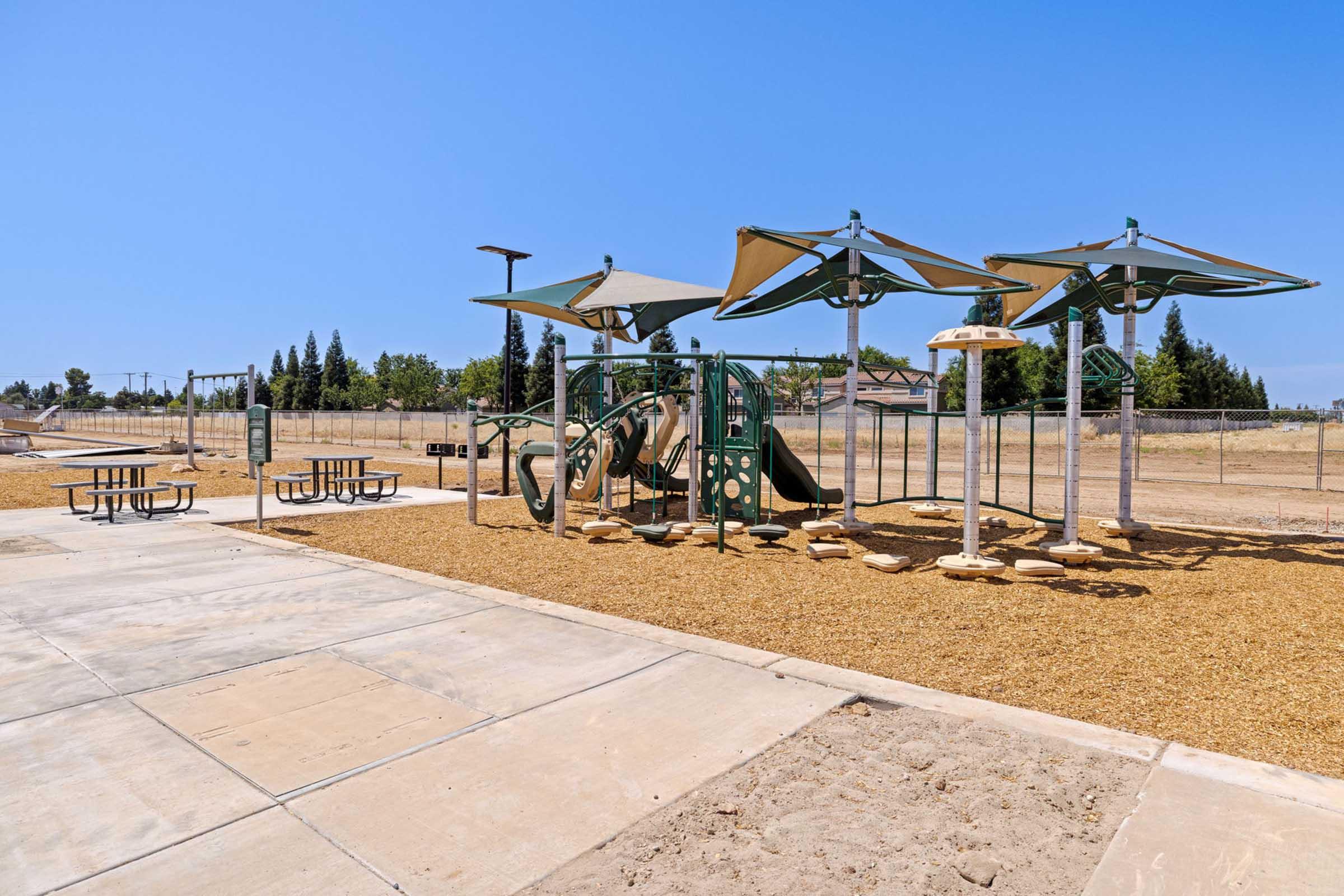 A playground featuring a slide, climbing structures, and shaded areas with large canopies. Nearby picnic tables are arranged on a sandy surface. The background shows a clear blue sky and trees, with a fenced area surrounding the playground.