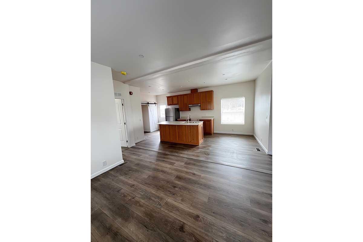 Spacious interior of a modern open-concept kitchen and living area. The room features light-colored walls, large windows, and solid wood flooring. A kitchen island with wooden cabinetry and stainless steel appliances is visible. The overall design is contemporary and well-lit.