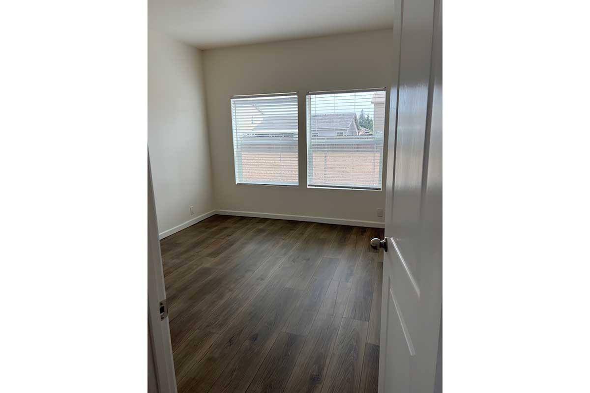 A clean, empty room with light-colored walls and wooden flooring. There are two windows with blinds allowing natural light to enter. A partially open white door is visible on the left side. The room is spacious and uncluttered, suitable for various furnishings.