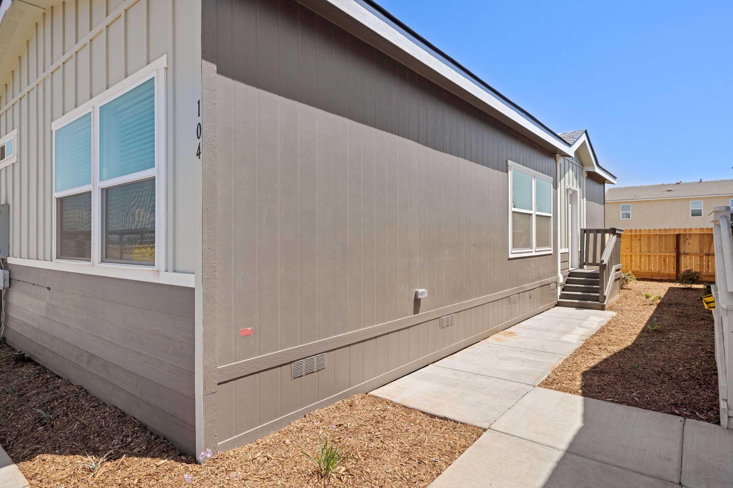 Side view of a single-story building with gray siding. A sidewalk runs alongside the structure, leading to a set of stairs at the back. There are small plants in the dirt and a wooden fence visible in the background, with a clear blue sky above.