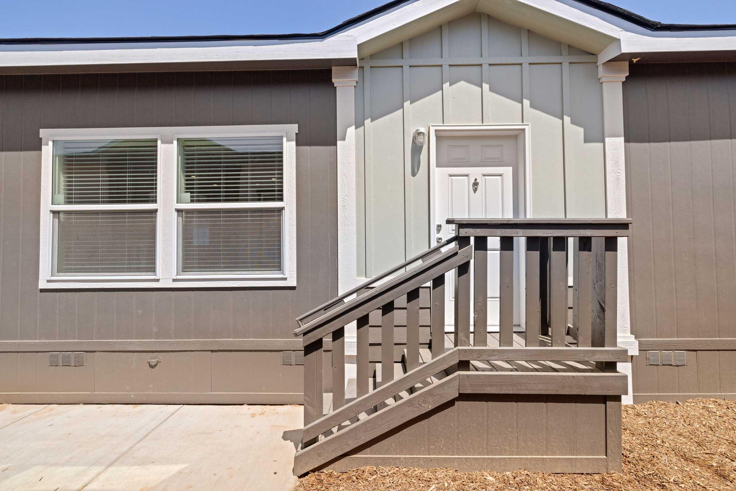A modern front entrance of a house featuring a white door with a simple light fixture above, flanked by two large windows. The exterior walls are a combination of light gray and dark gray siding, and there is a wooden ramp leading up to the door, set against a gravel surface. The sky is clear and blue.