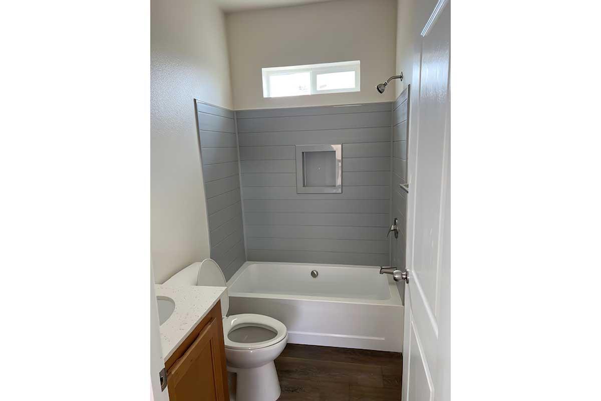 A modern bathroom featuring a white bathtub with a shower, a wall with gray paneling, and a white toilet. There is a small window above the bathtub and a sink area with a light-colored countertop. The flooring appears to be dark wood. The door is white, matching the overall clean aesthetic.
