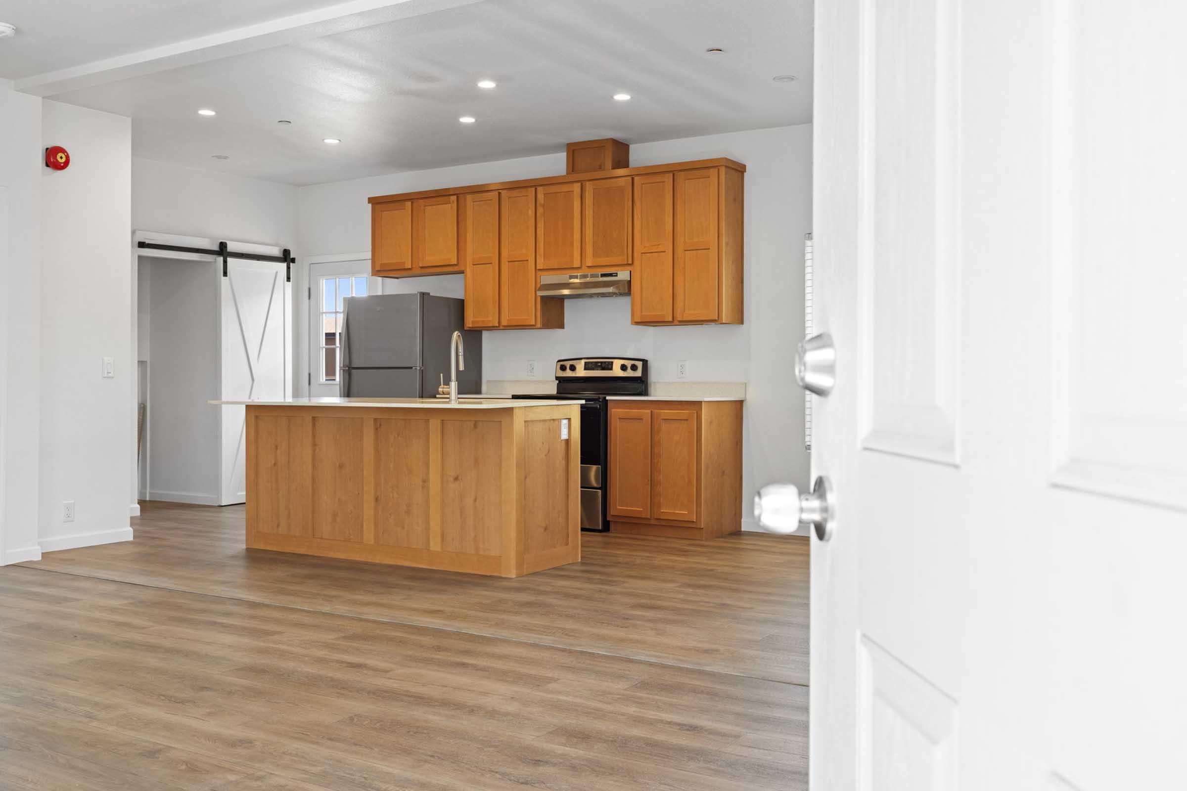 A modern kitchen with brown wooden cabinets and a large island featuring a countertop. Stainless steel appliances, including a refrigerator and gas stove, are visible. The flooring is light wood, and the space is bright with recessed lighting and a door partially open in the foreground.
