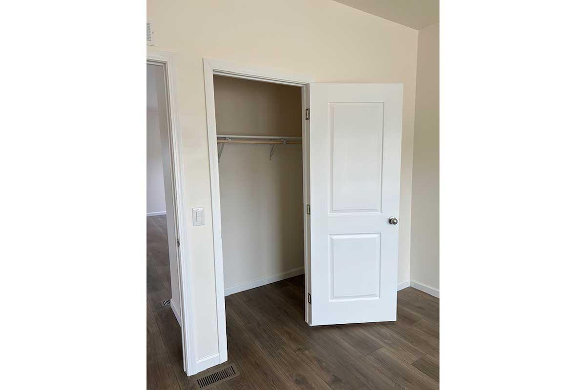 A partially open white closet door in a light-colored room with neutral walls and wooden flooring. The closet interior is empty, featuring a hanging rod. The adjacent wall has a light switch, and there is another doorway visible in the background.