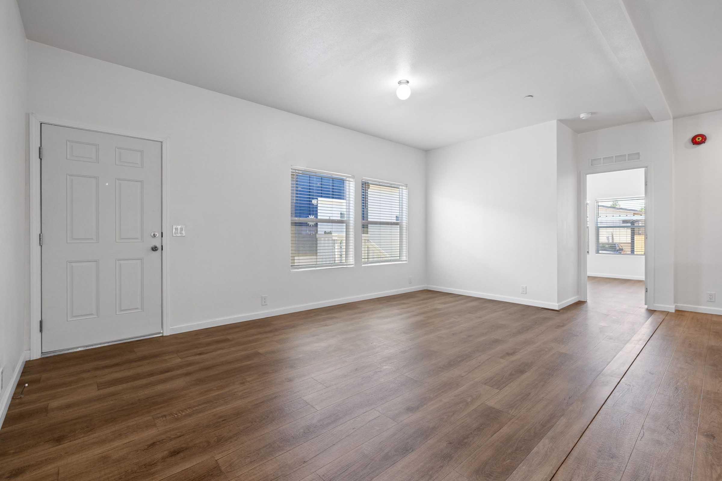 A bright, empty room featuring light-colored walls, large windows, and a wooden floor. The entrance door is on the left, with another room visible through an open door on the right. Natural light illuminates the space, creating a welcoming atmosphere.