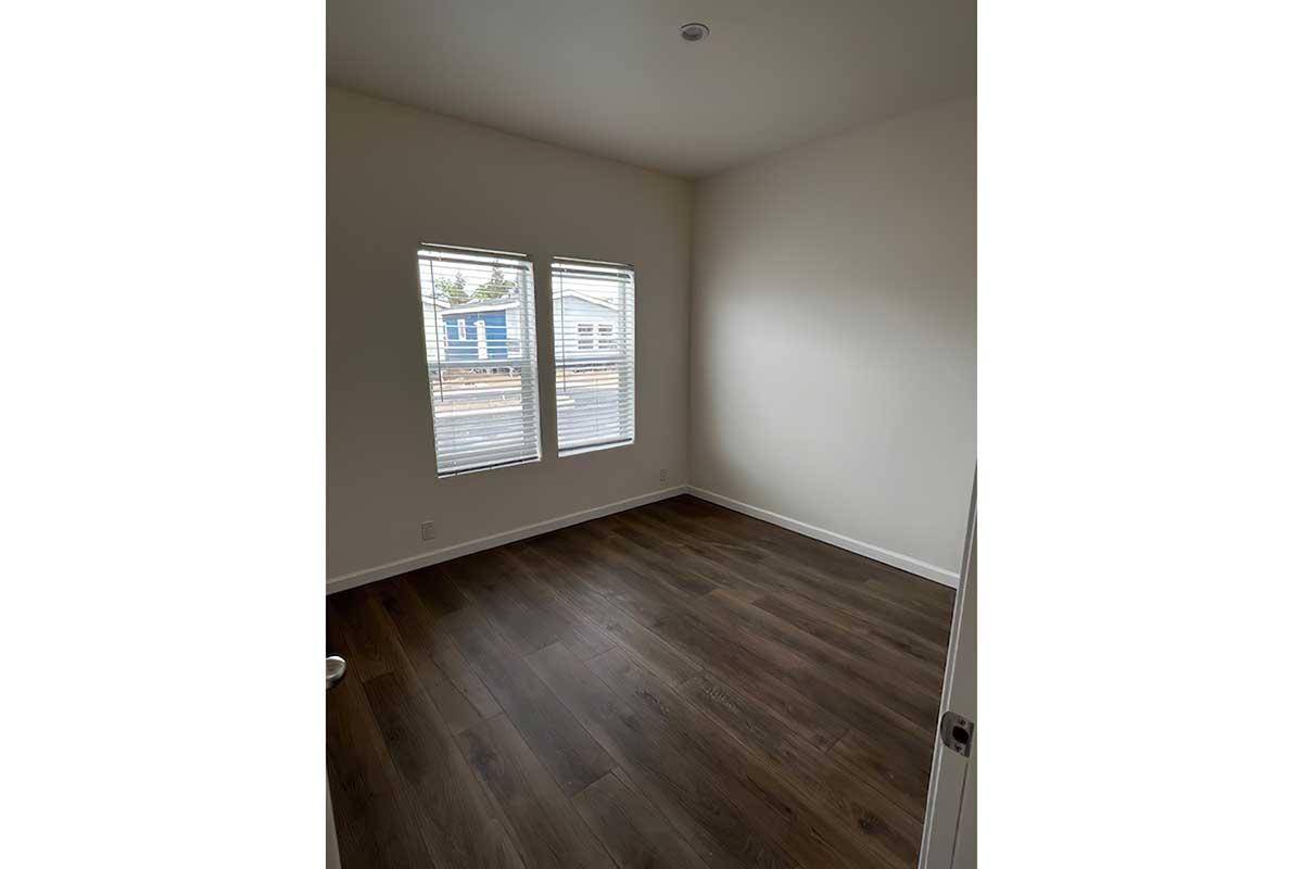 A small, empty room with light-colored walls and two windows. The flooring is a dark wood, and there is a door visible on the left. Natural light enters through the windows, illuminating the simple, uncluttered space, which has a plain and minimalist appearance.