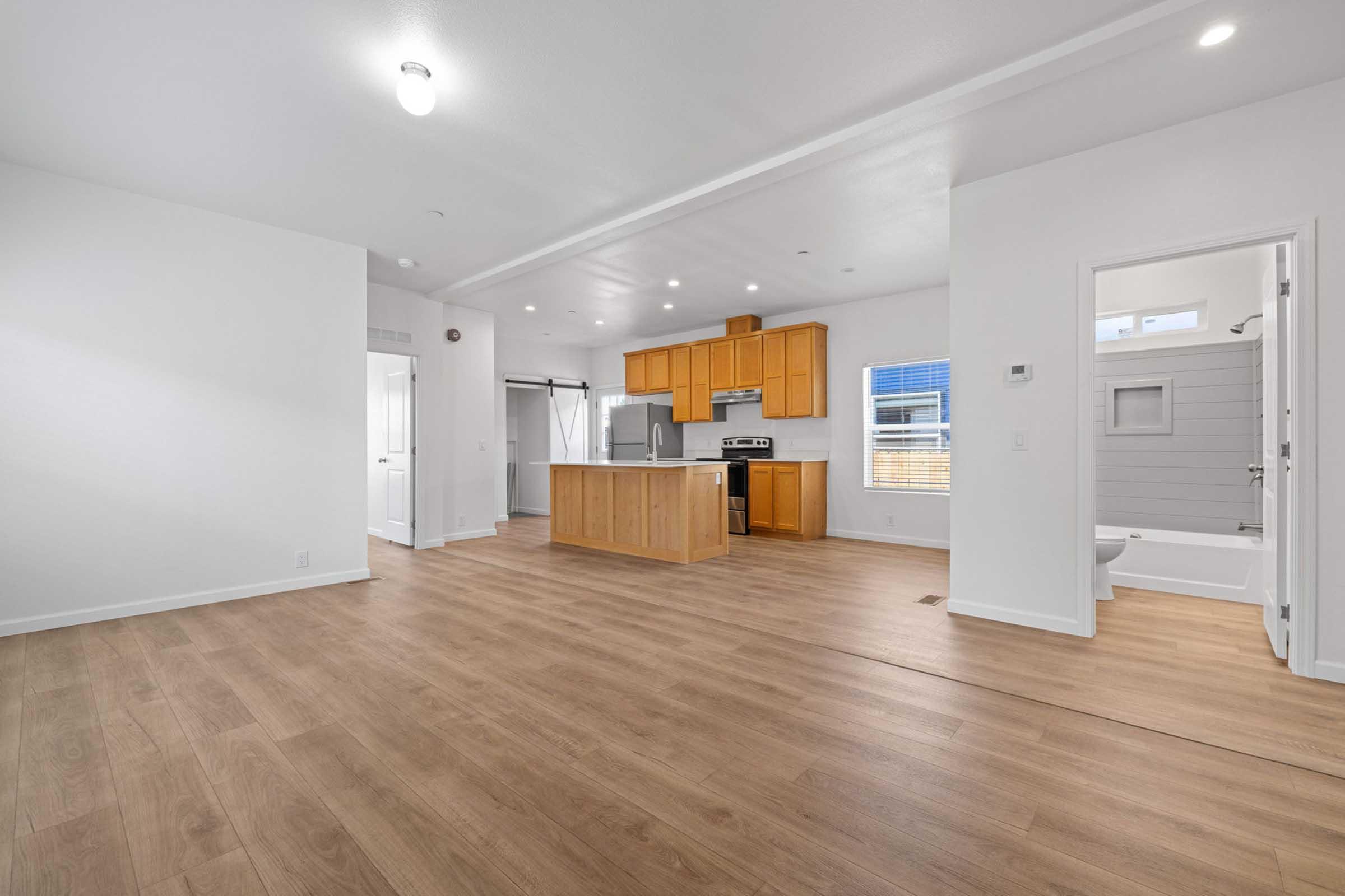 Interior view of a modern, open-concept living space featuring a kitchen with wooden cabinets, an island, and stainless steel appliances. The floor is light-colored wood, and there are large windows allowing natural light. A door leads to a bathroom, and the walls are painted in a clean, white finish.