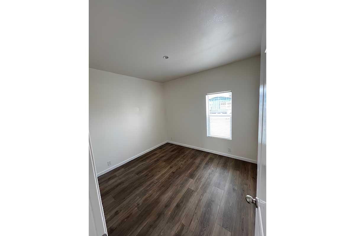Empty room with light-colored walls and hardwood flooring. A single window allows natural light to enter, creating a bright atmosphere. The room is unfurnished, emphasizing the spaciousness and neutral tones.