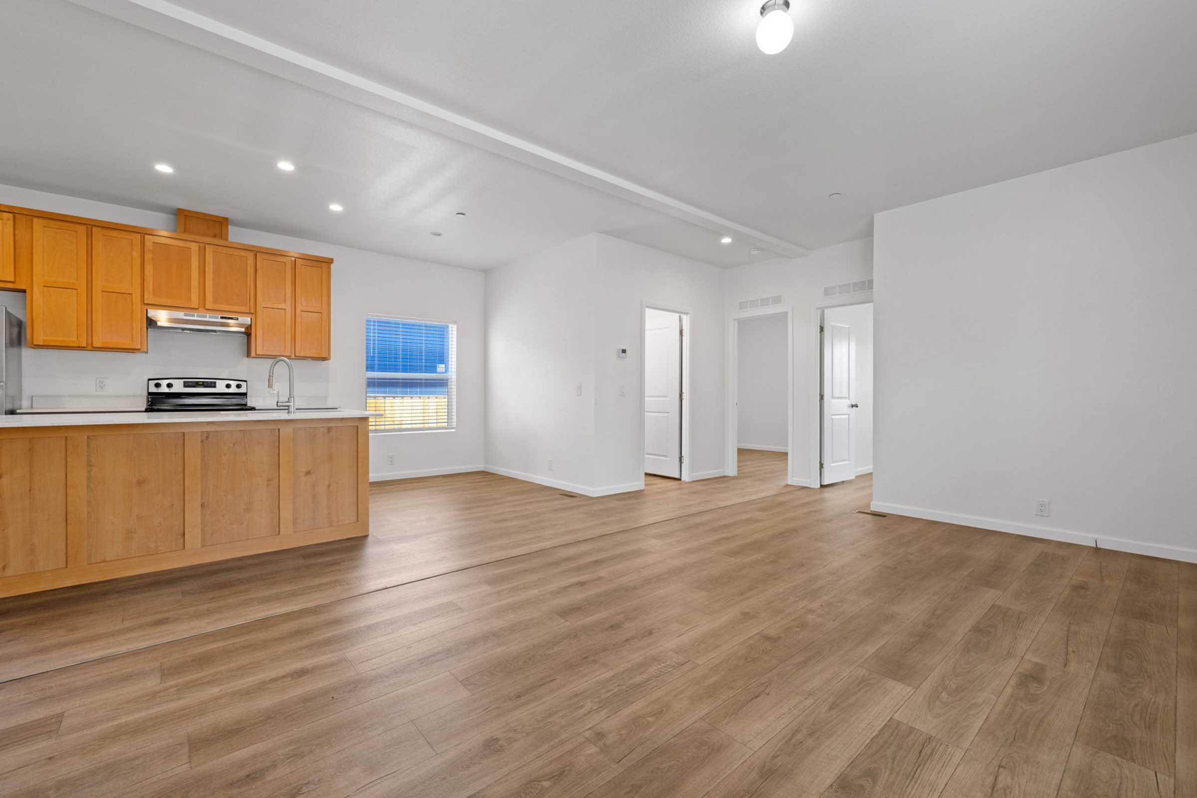 A modern open-concept living space featuring a kitchen with wooden cabinets, a stainless steel stove, and a window providing natural light. The flooring is light wood, and there are white walls. A hallway leads to additional rooms, creating a spacious and inviting atmosphere.