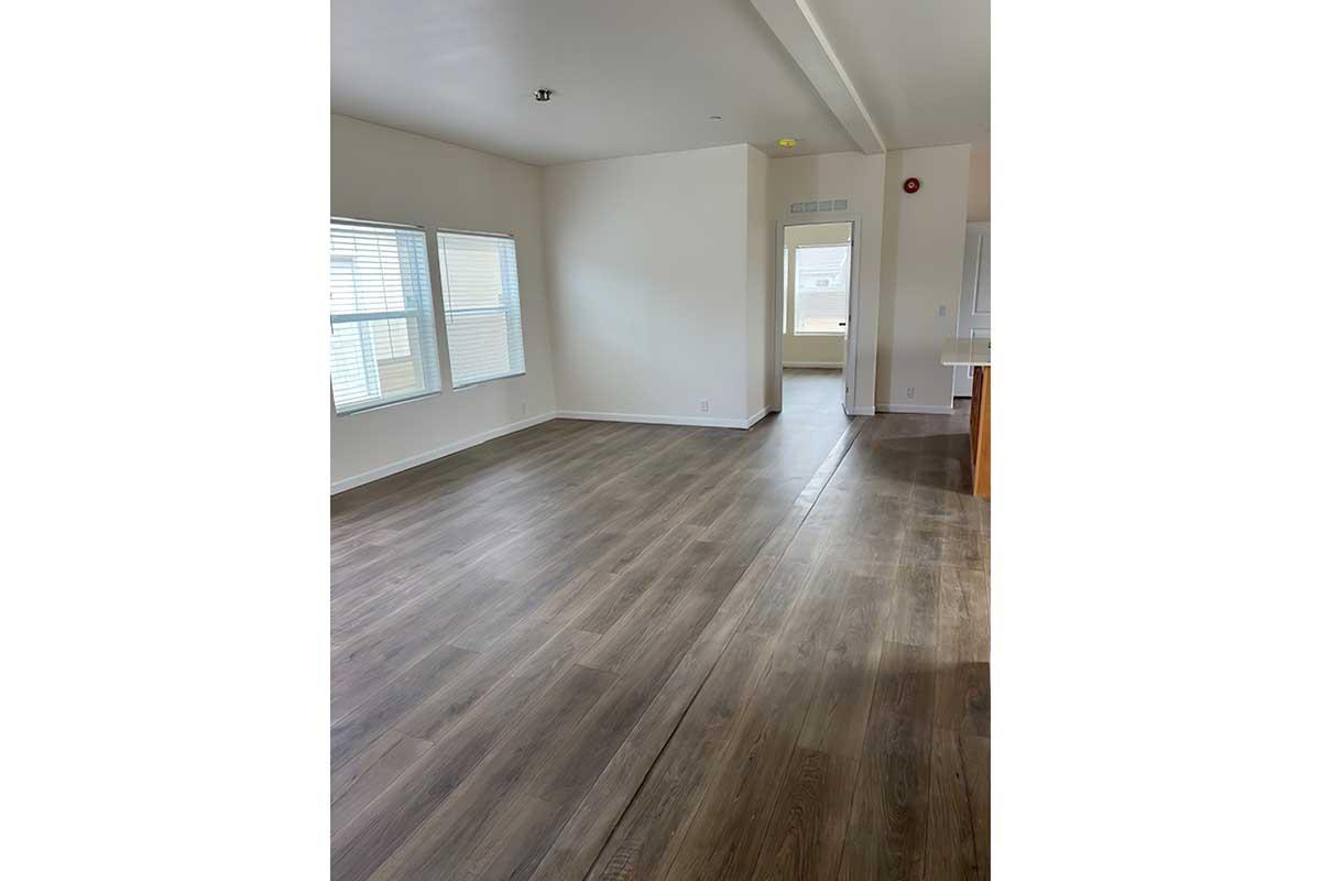 A spacious, empty room with light-colored walls and large windows allowing natural light. The floor is made of light brown wood laminate. In the background, there is an entryway leading to another area of the home. The overall ambiance is bright and airy.