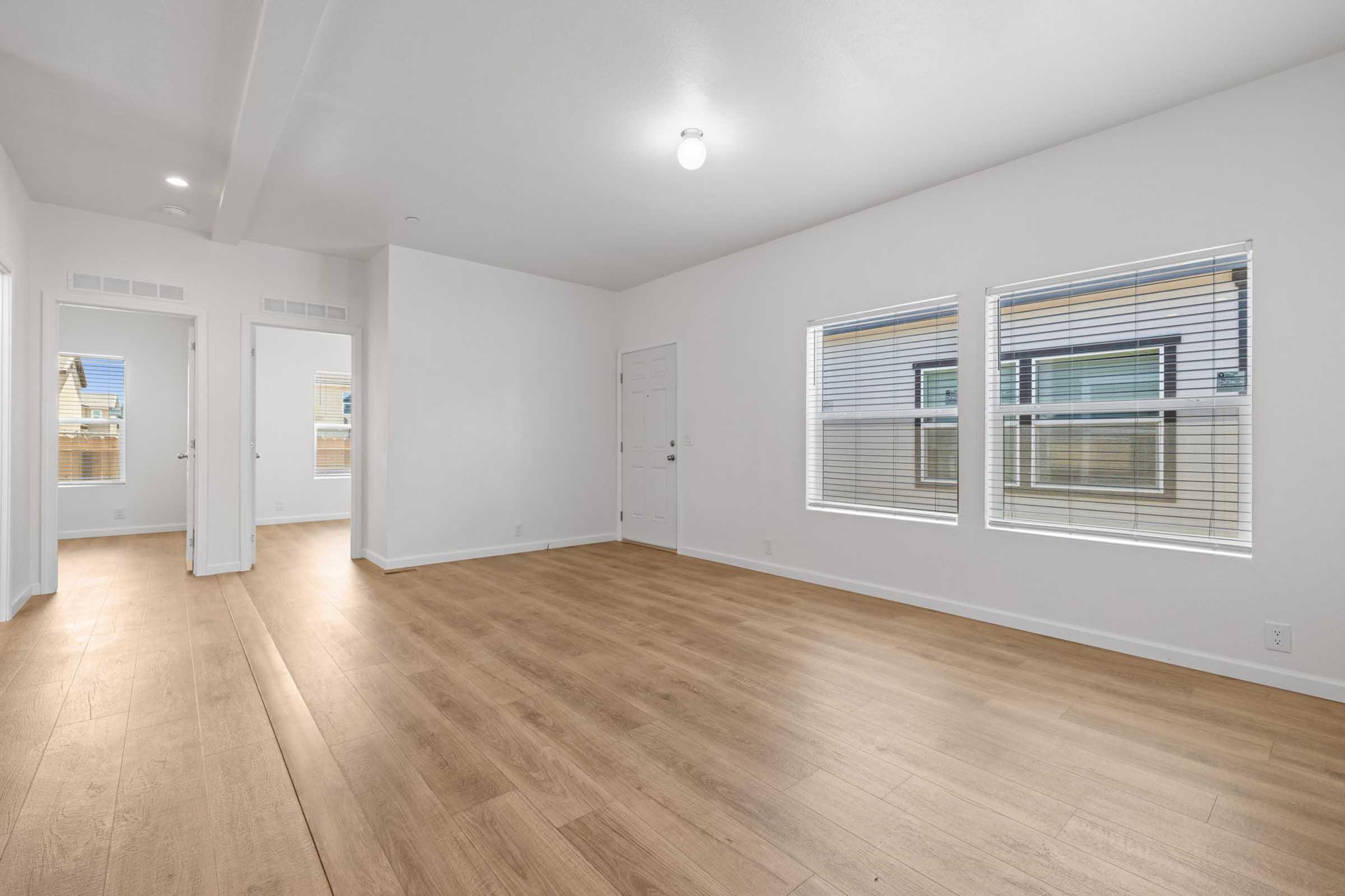 A bright, empty room featuring light-colored walls and hardwood flooring. There are two windows allowing natural light to enter, and a doorway leading to another room is visible in the background. The overall atmosphere is clean and modern, with a minimalistic design.