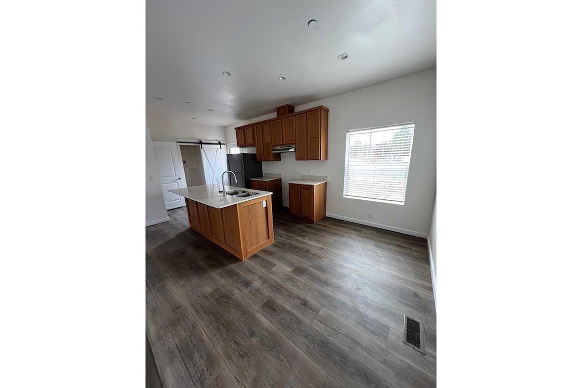 Modern kitchen featuring wooden cabinets, a large island with a sink, and stainless steel appliances. The space has gray wood-like flooring and ample natural light from a window. A sliding barn door is visible on one side, and the overall color scheme is warm and inviting.