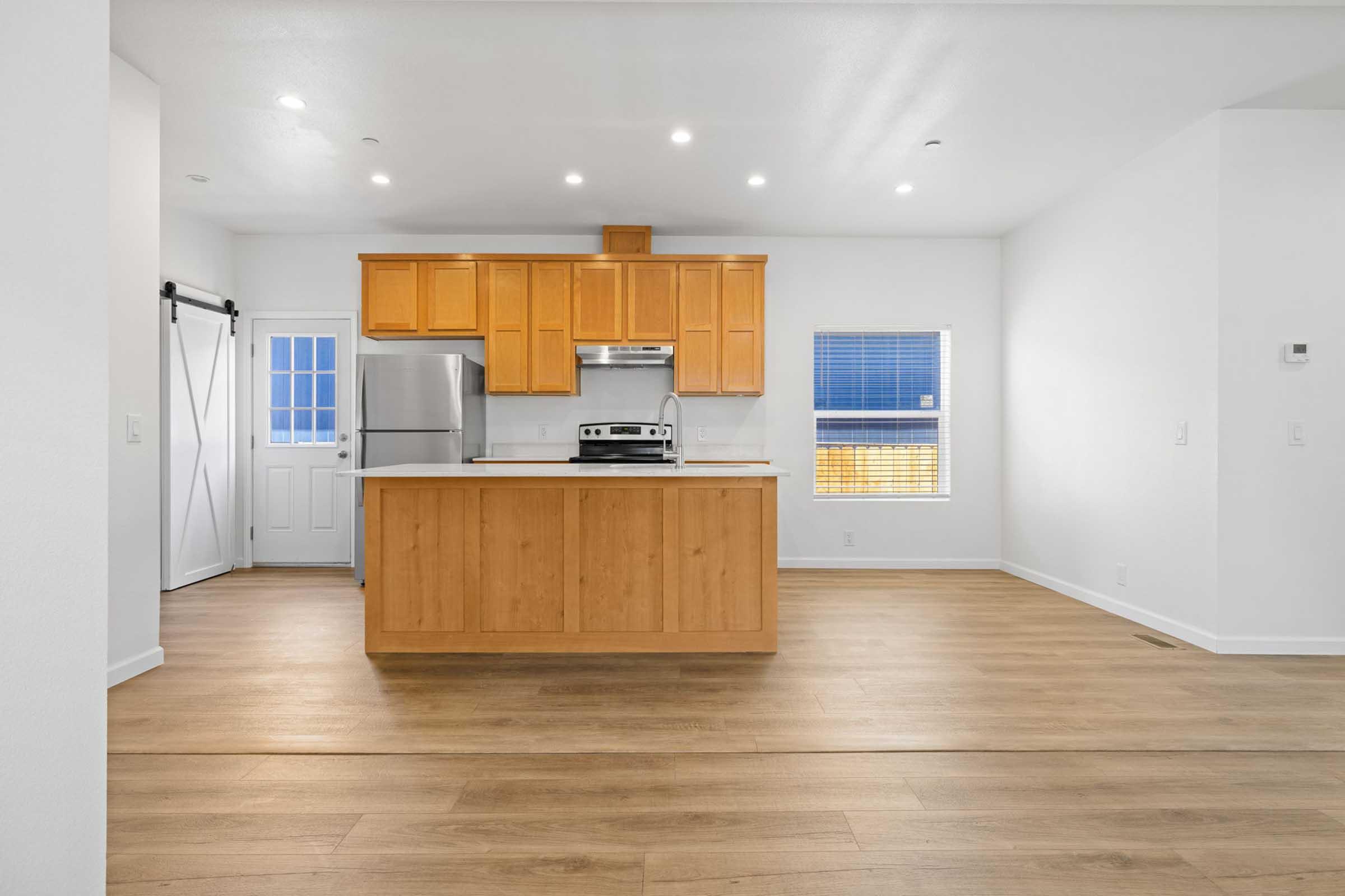 Modern kitchen with wooden cabinetry, stainless steel appliances, and a large central island. The space features open layout with natural light coming from a window and door. Light-colored walls and hardwood flooring create a bright and inviting atmosphere.