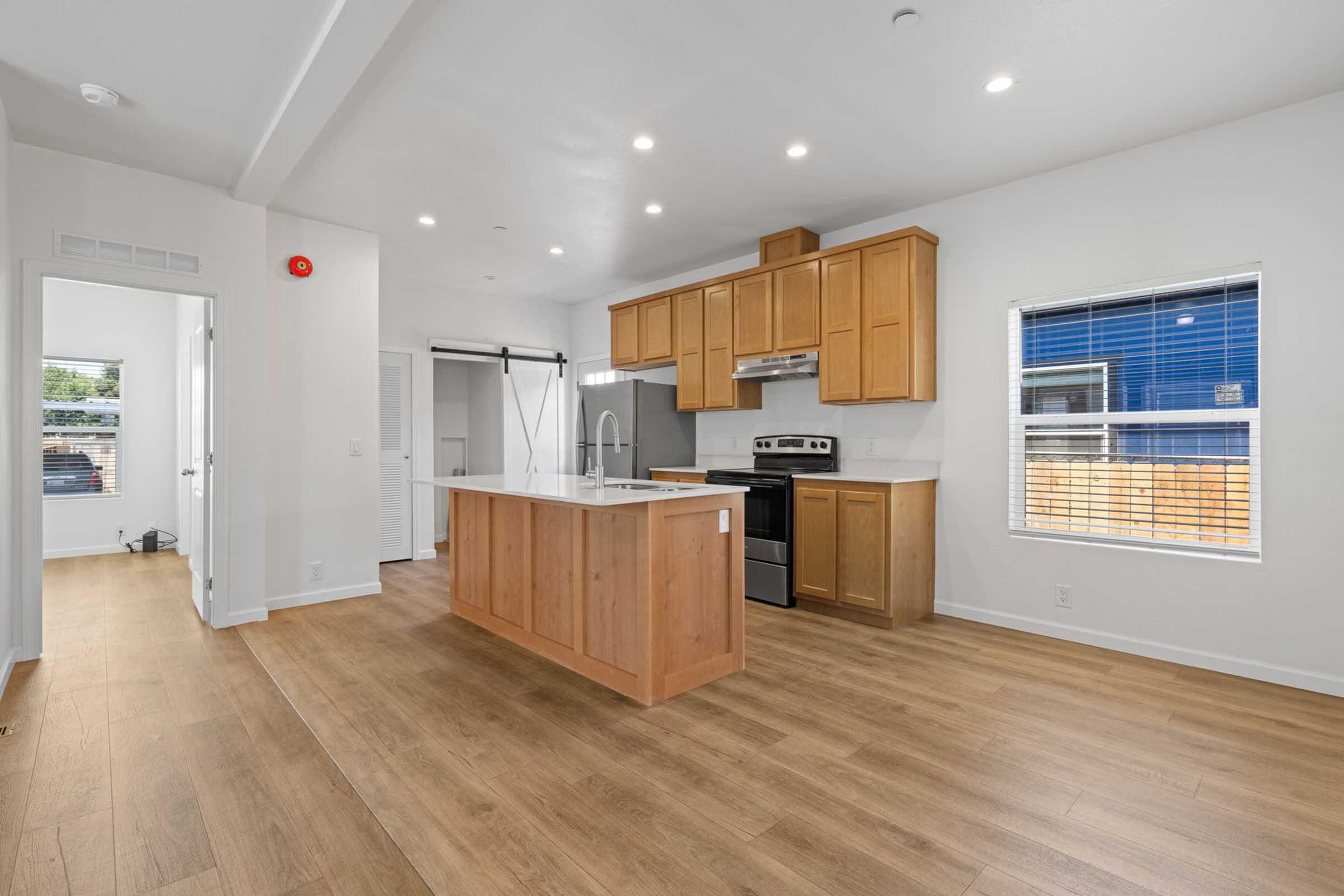Bright and modern interior of a kitchen featuring wooden cabinets, a stainless steel stove, and an island with a countertop. Large windows allow natural light to fill the space, highlighting the light-colored walls and wood-look flooring. A doorway leads to another room, providing a glimpse of an open layout.