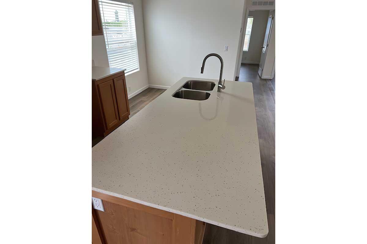 A modern kitchen countertop with a smooth white surface featuring speckles. It includes a double sink and a sleek faucet, with cabinetry visible in the background. The space is well-lit by natural light coming through nearby windows, highlighting the clean and contemporary design.