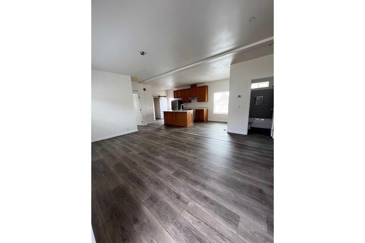 Spacious interior of a modern apartment featuring light wood laminate flooring, white walls, and a kitchen area with wooden cabinetry. Large windows provide natural light. The layout includes a living space with an open-plan design leading to the kitchen.