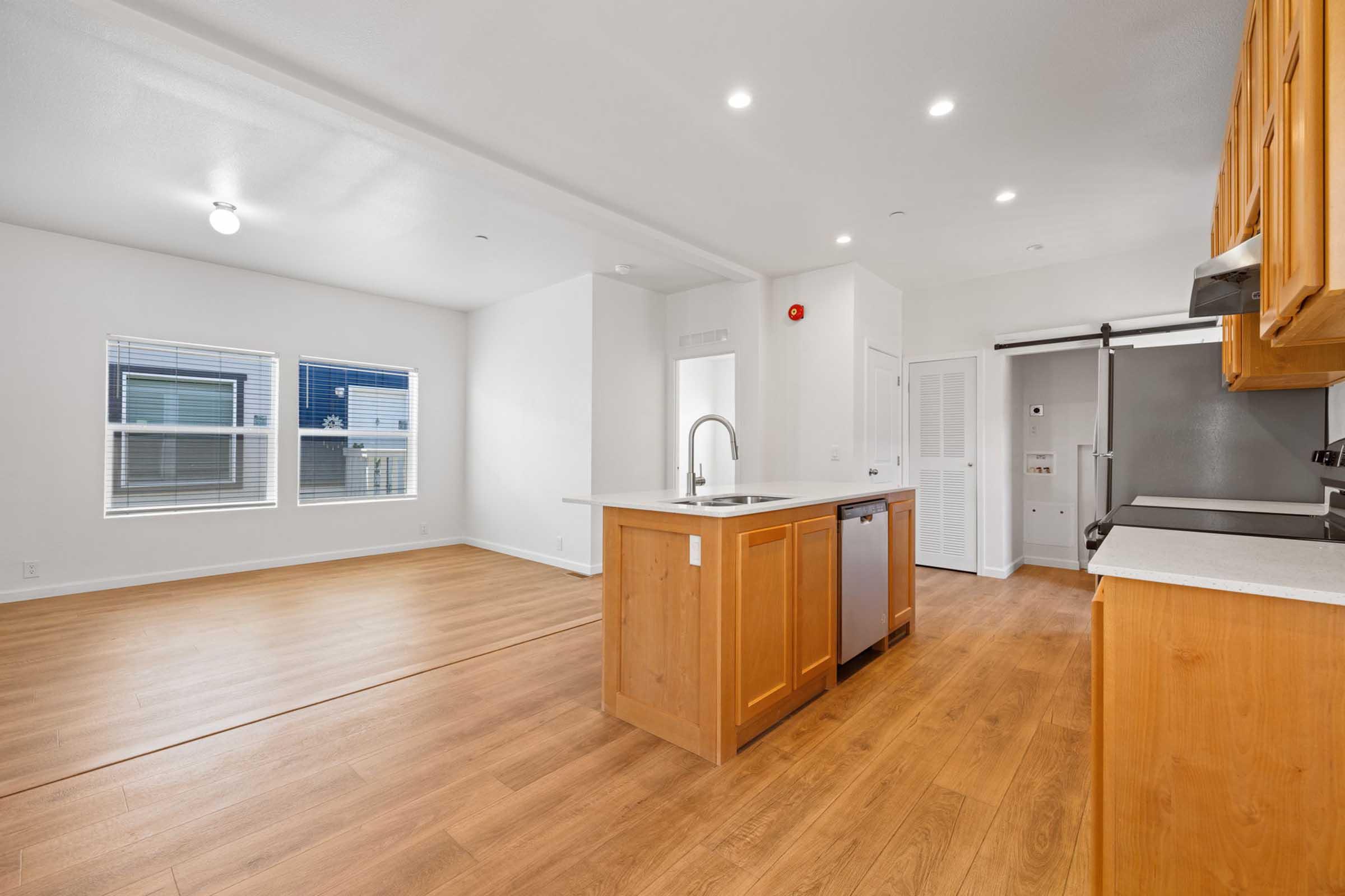 Modern kitchen and living area featuring wooden cabinets, an island with a countertop, stainless steel appliances, and large windows providing natural light. The flooring is light wood, and the walls are painted white, giving the space a bright and open feel.