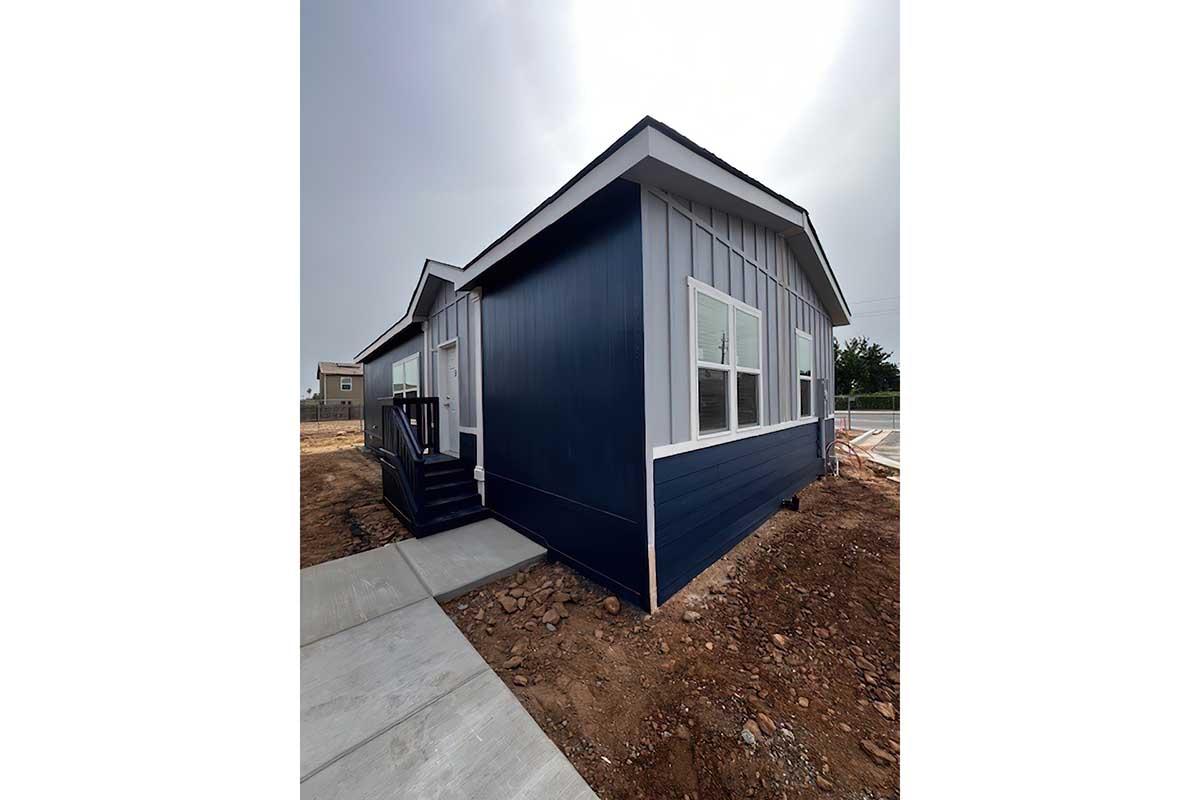 A new single-story blue house with white trim, featuring large windows and a porch. The house is situated on a construction site with dirt and gravel around it, and a concrete pathway leading up to the entrance. The sky appears overcast, indicating cloudy weather.