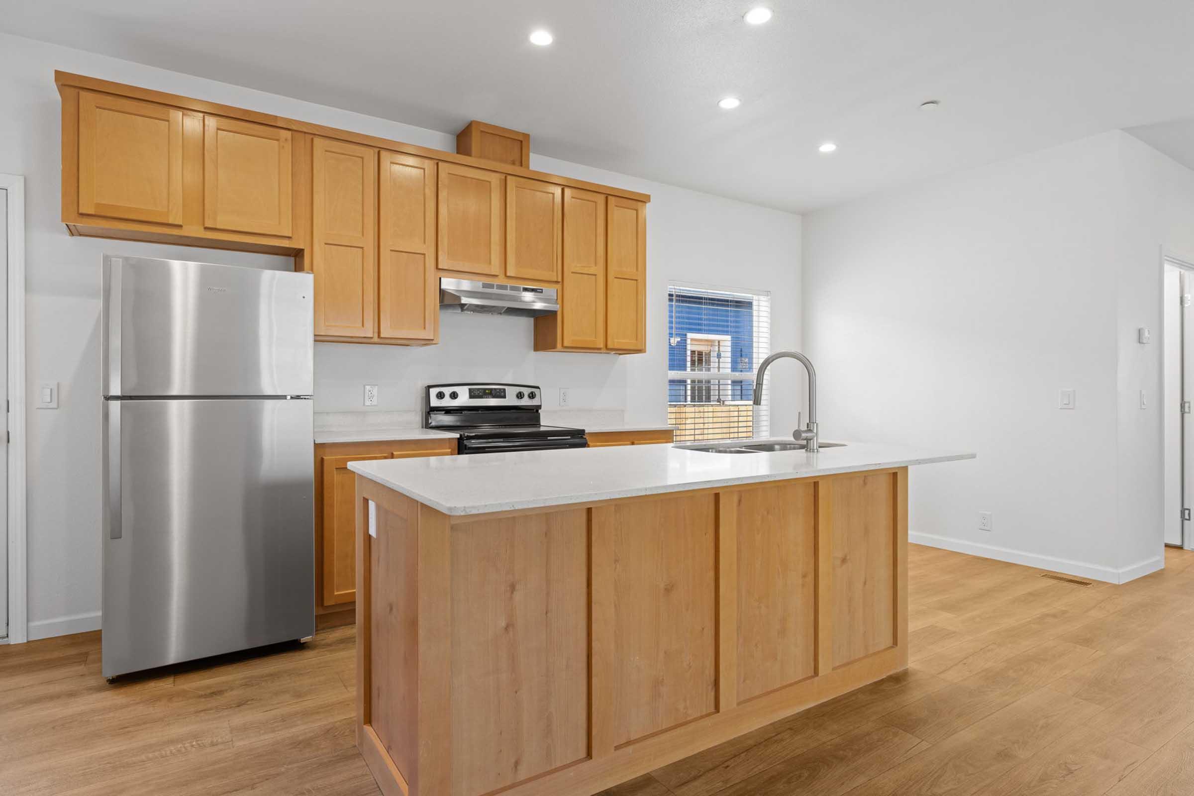 Modern kitchen featuring light wood cabinetry, a stainless steel refrigerator, and a sleek stove. An island with a countertop provides additional workspace. The floor is made of light wood, and there are recessed ceiling lights for bright illumination. A window allows natural light to enter the space.