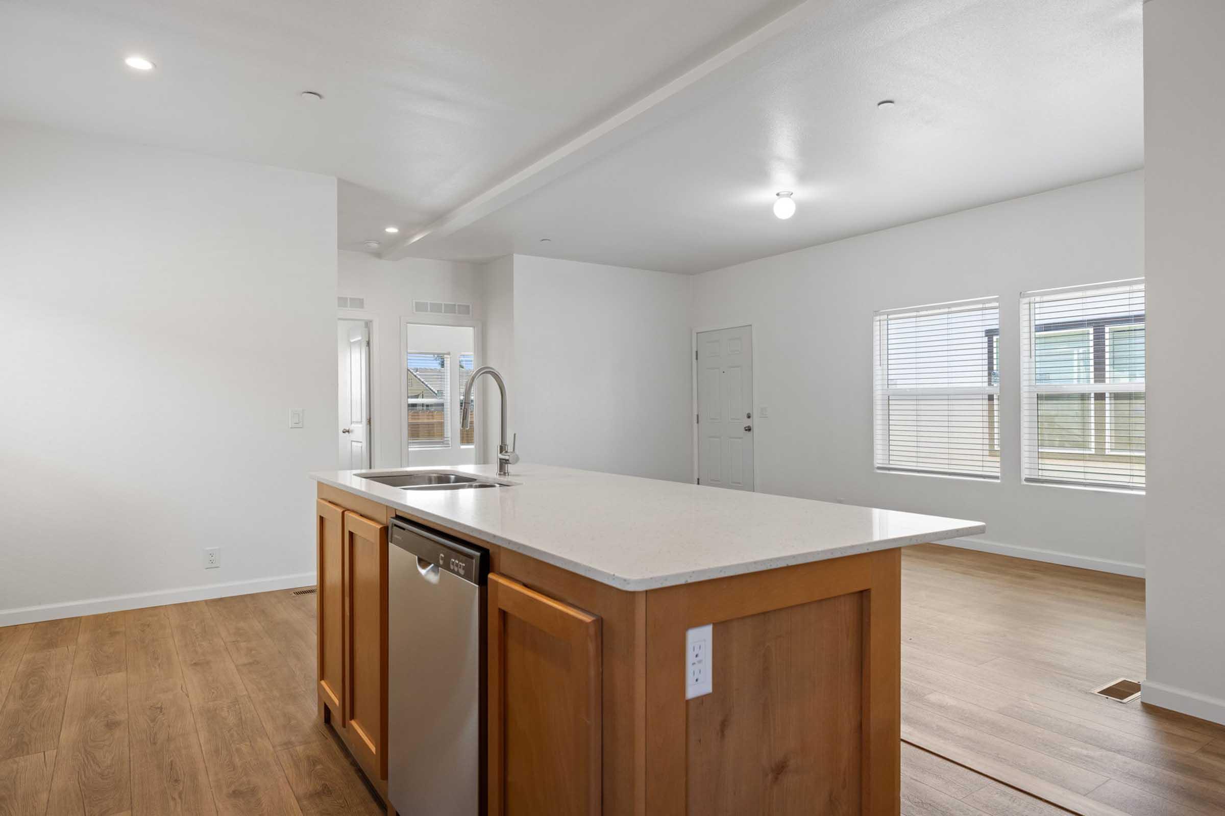 A modern kitchen area featuring an island with a sink and dishwasher, surrounded by light-colored cabinets. The open layout leads to a bright living space with large windows. The flooring is light wood, and the walls are painted white, creating a fresh and airy atmosphere.