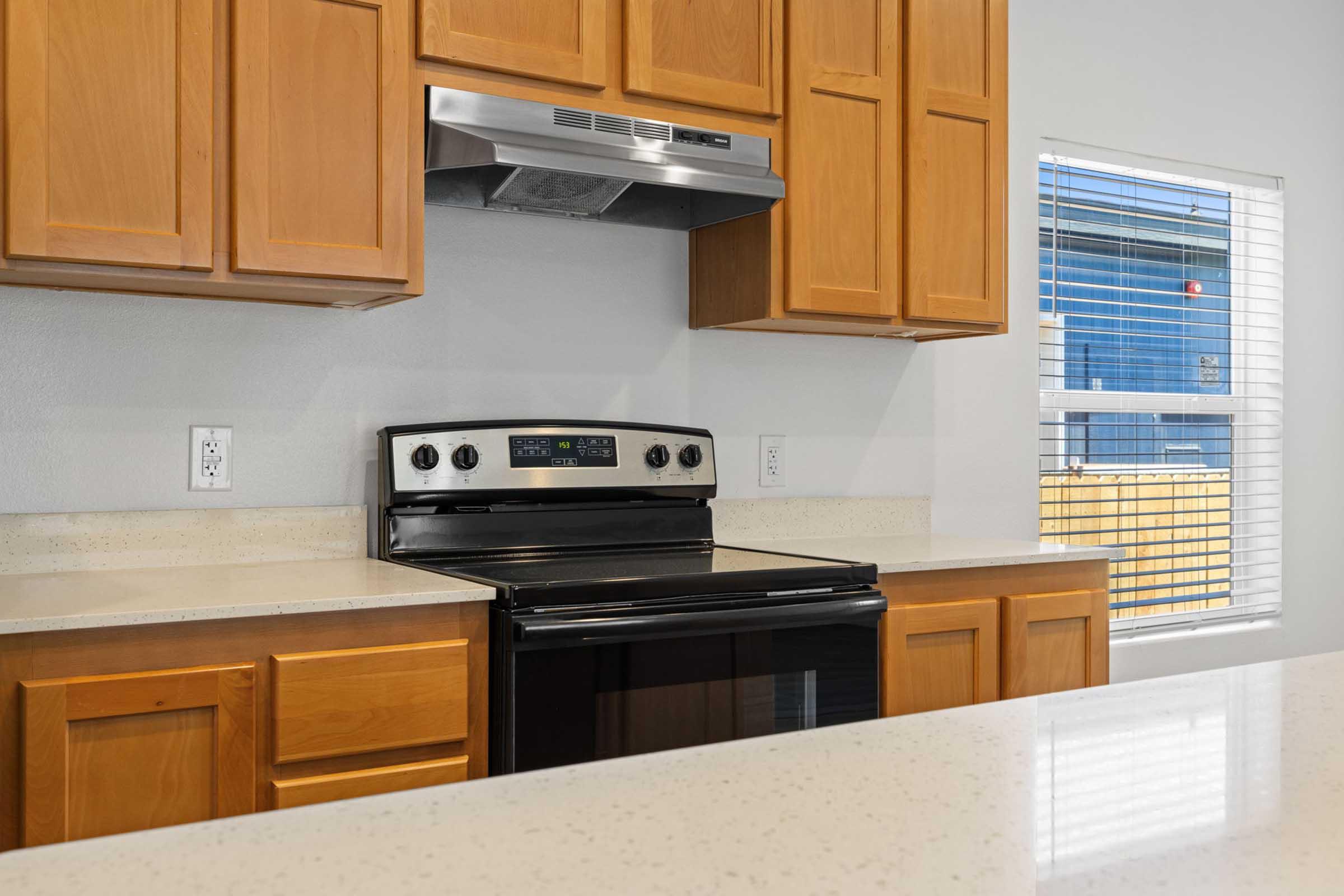 A modern kitchen featuring an electric range with an oven, a stainless steel hood above, wooden cabinets, and a light-colored countertop. A window with blinds provides natural light, overlooking an outdoor area.