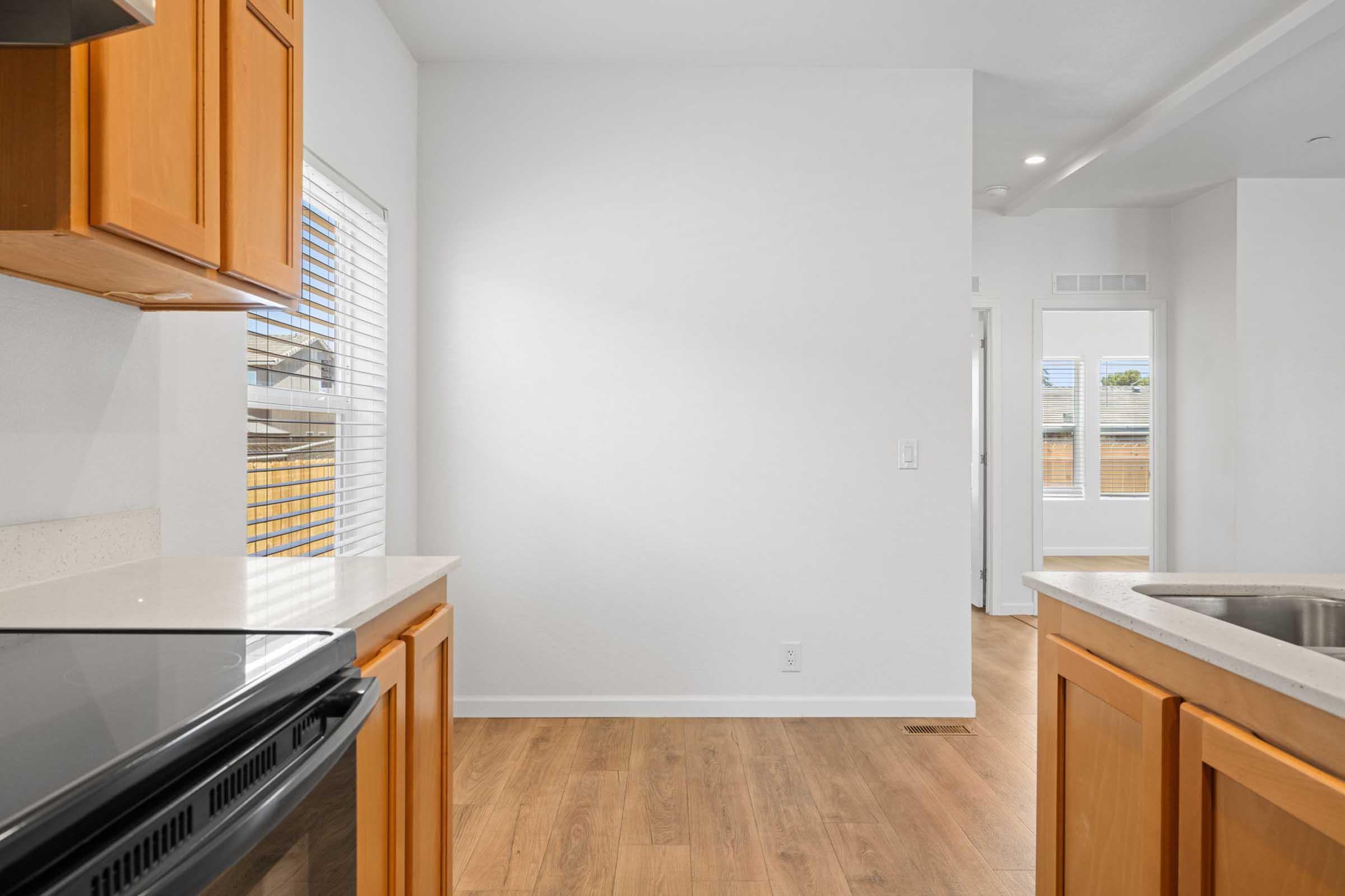 A modern kitchen featuring wooden cabinets, a black stove, and a countertop with a sink. The room is brightly lit with large windows allowing natural light to enter, and hardwood flooring extends into the adjacent living area. The walls are painted white, creating a clean and open atmosphere.