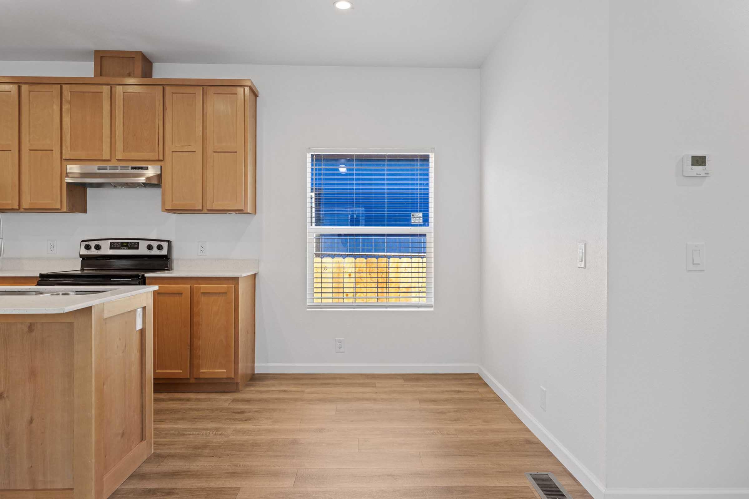 A modern kitchen featuring wooden cabinetry, a black stove, and a window with blue outdoor view. The room has light-colored walls and flooring, creating a spacious and bright atmosphere. A thermostat is visible on the wall, and the layout includes a central island.