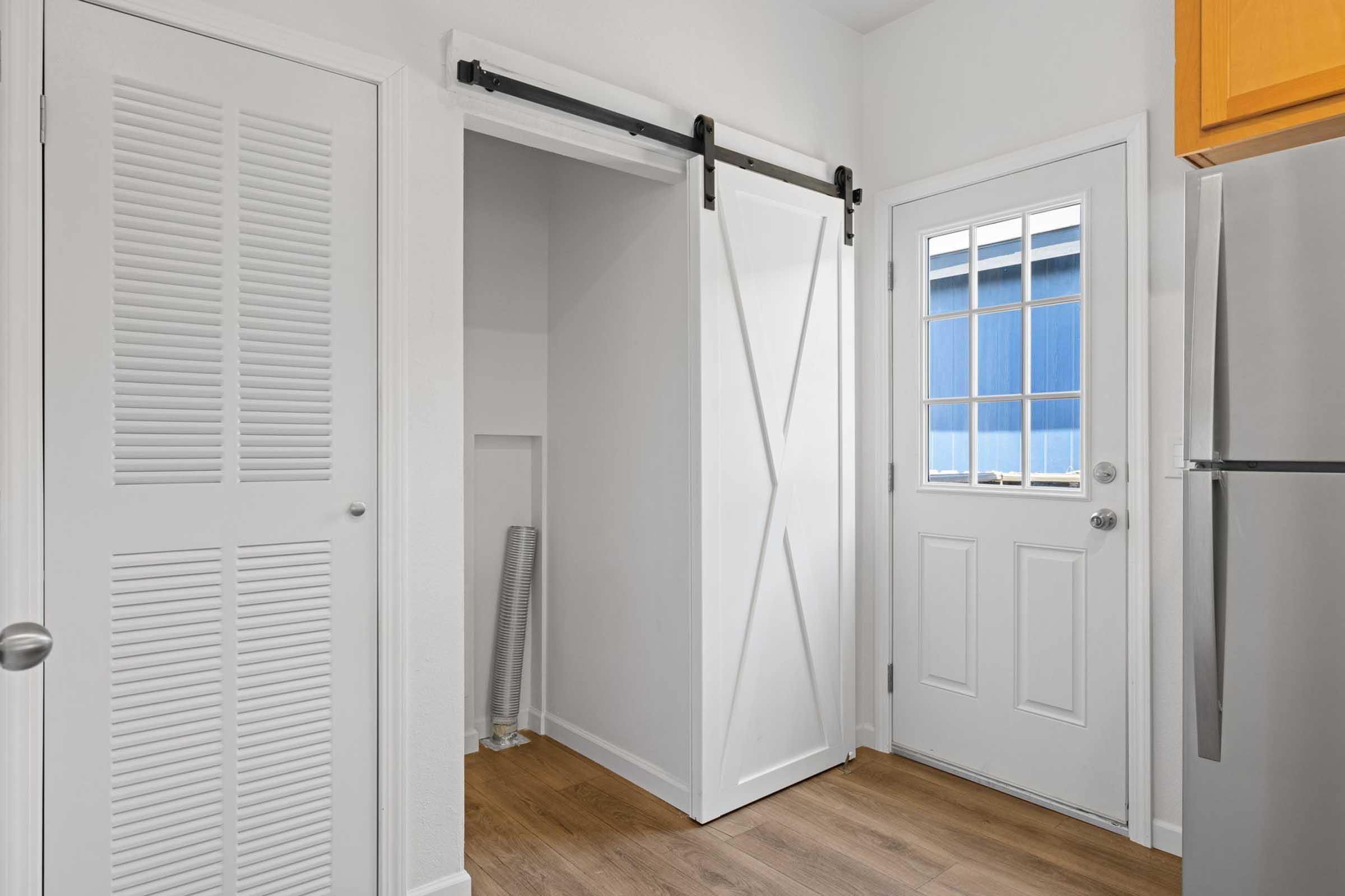 Interior view of a bright, modern entryway featuring a white sliding barn door, a closet with louvered doors, and a door with window panes leading outside. Light wood flooring and a stainless steel fridge are visible in the background, creating a clean and welcoming atmosphere.