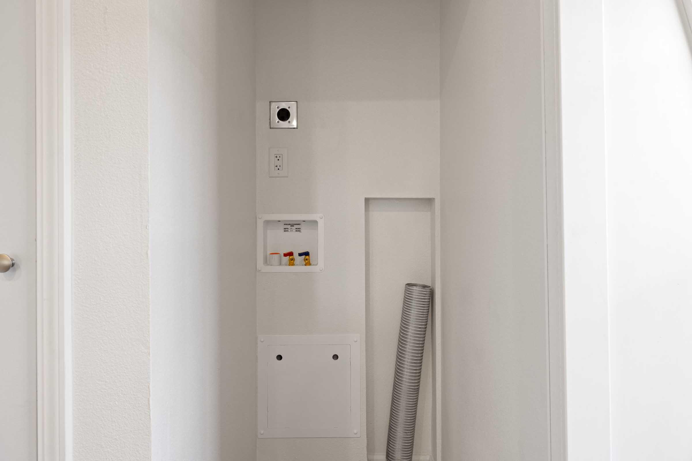 A small, minimalistic utility space featuring a white wall with a vent, electrical outlet, and an electrical panel. There is a recessed area in the wall with a blank cover and a flexible grey duct lying on the floor. The overall color scheme is light and clean, emphasizing simplicity.