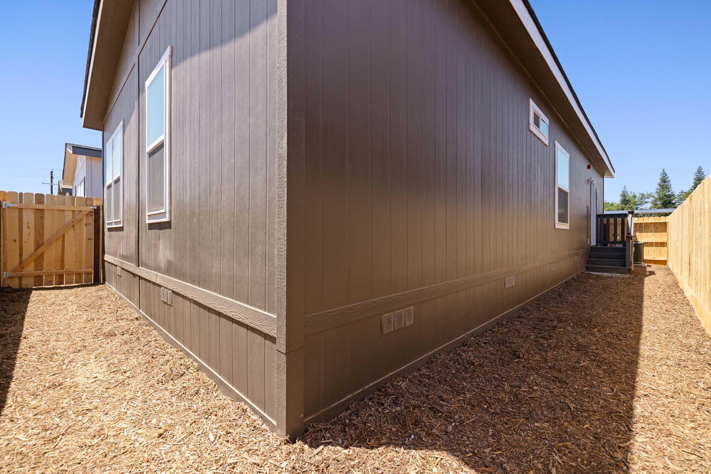 The image shows the side view of a brown-colored house with several windows. There is a wooden fence adjacent to the house, and the ground is covered with mulch. The sky is clear and blue, providing a bright backdrop to the scene. The angle emphasizes the house's corner and the surrounding outdoor area.