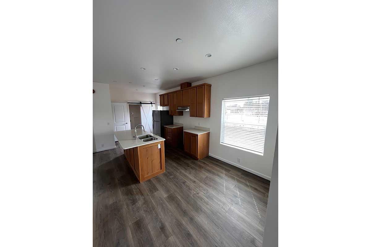 A modern kitchen with wooden cabinets, a central island, and a sink. Large window providing natural light, and a view of an adjacent room. The floor is dark laminate, and the walls are painted white. The space appears clean and well-organized.