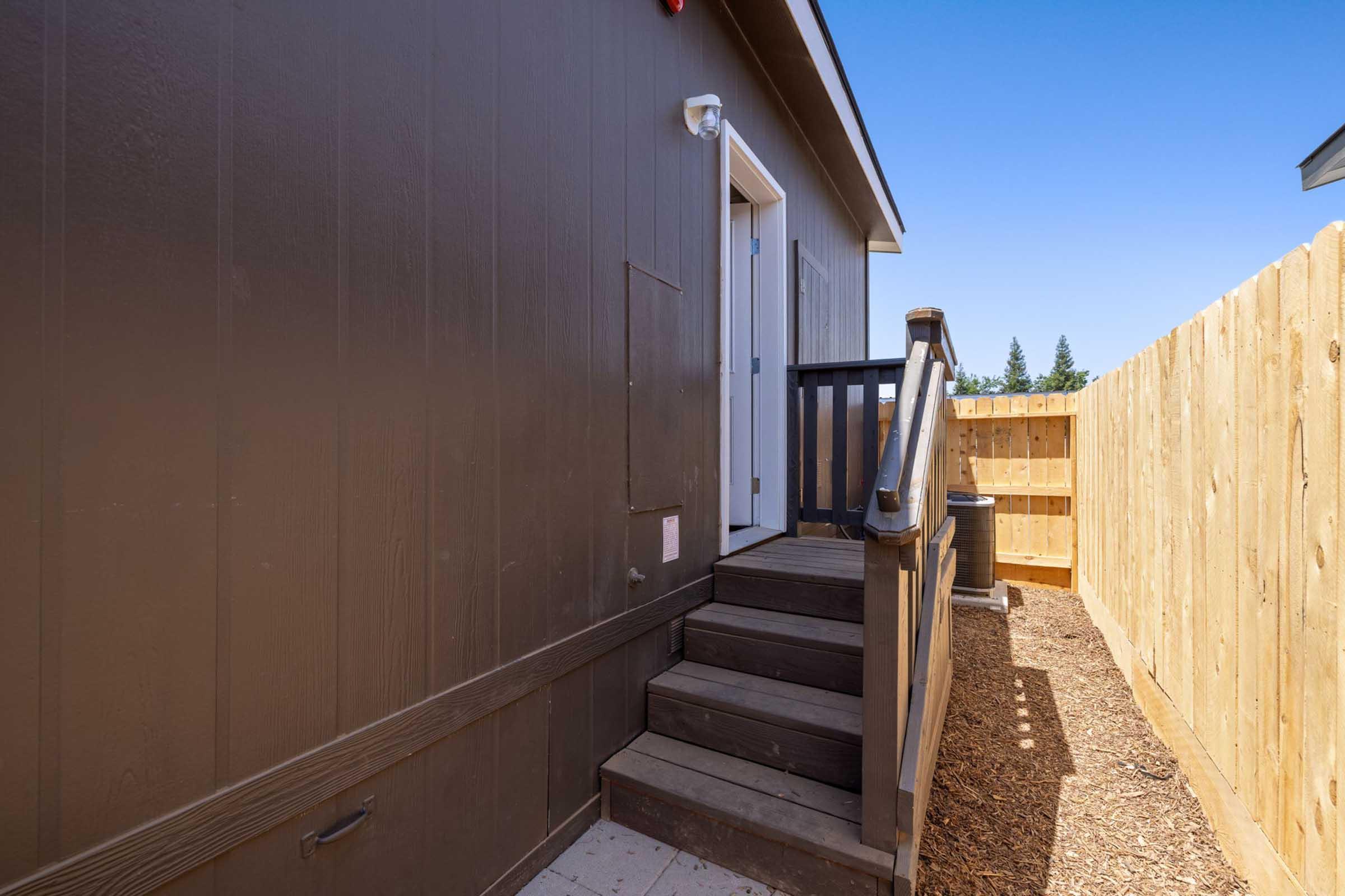A wooden staircase leading to a door on the side of a brown building, surrounded by a wooden fence, with clear blue skies in the background. The area features gravel flooring and a well-maintained appearance.