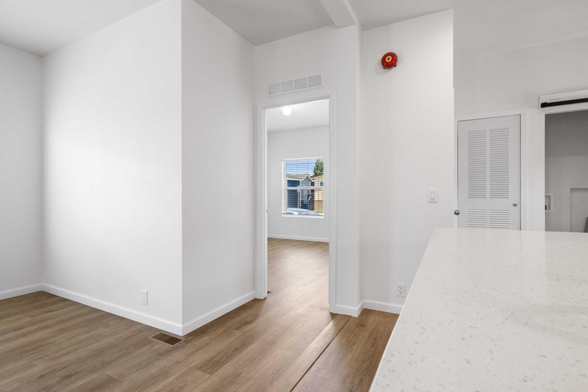 A modern, airy interior space featuring light-colored walls and wood flooring. The image shows an open layout with a view of a doorway leading to another room, brightened by natural light coming through a window. The room has minimal decor and a clean, minimalist design.