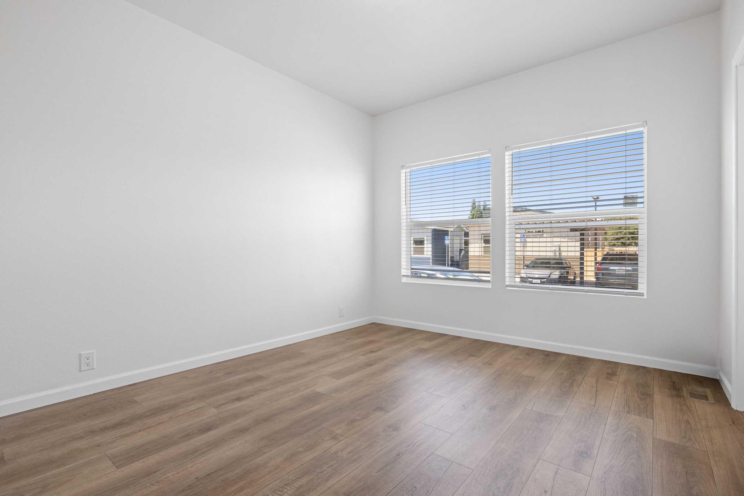 Bright, empty room featuring light wood flooring and two large windows with white blinds. The walls are painted a soft white, creating a clean and spacious feel. Natural light floods the space, highlighting the simplicity and openness of the area. Ideal for potential use as a living room or office.