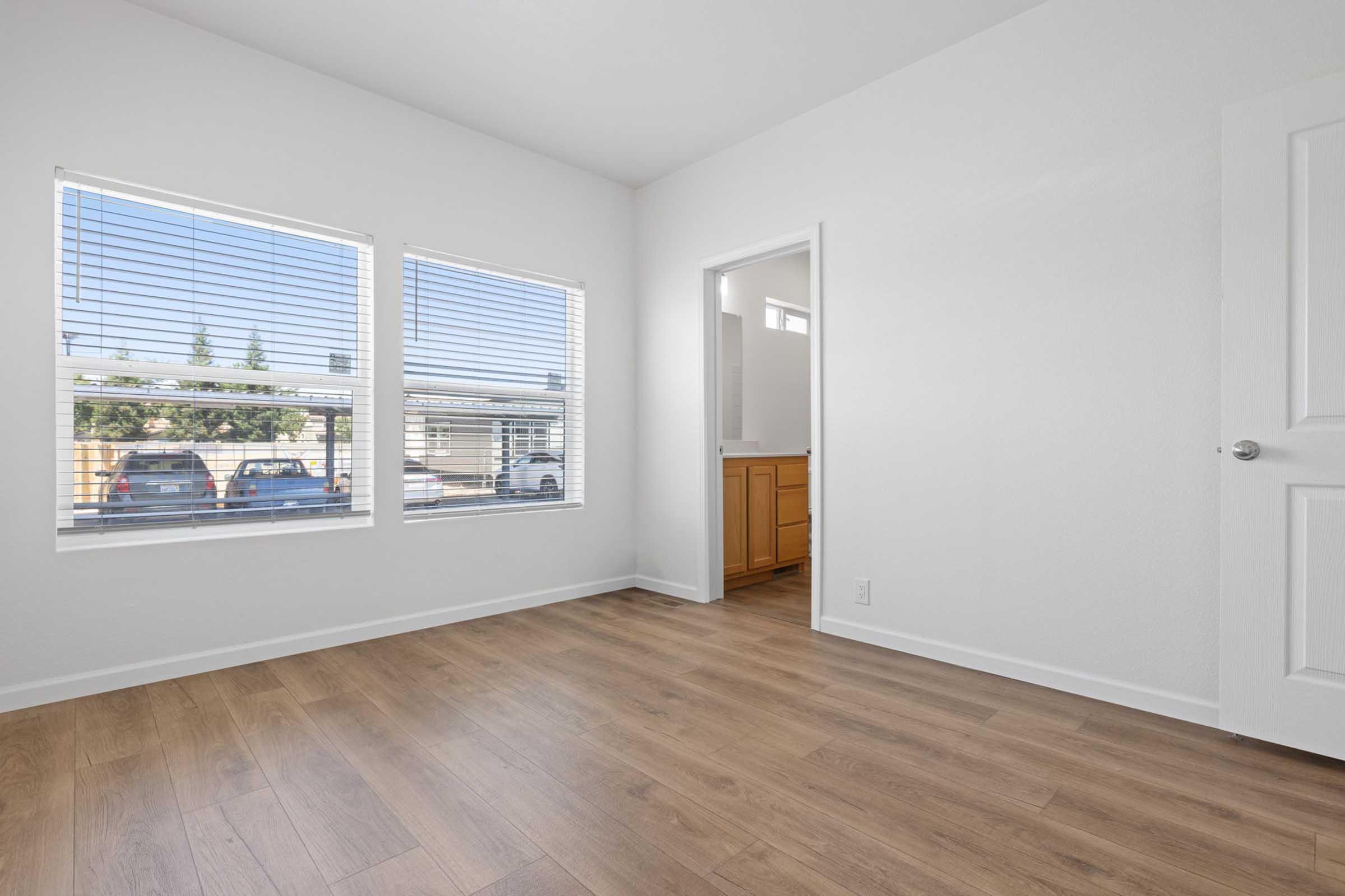 A bright, empty room with hardwood flooring, featuring two large windows with white blinds that let in natural light. A doorway leads to another room with wooden cabinetry visible. The walls are painted white, creating a clean and spacious atmosphere.