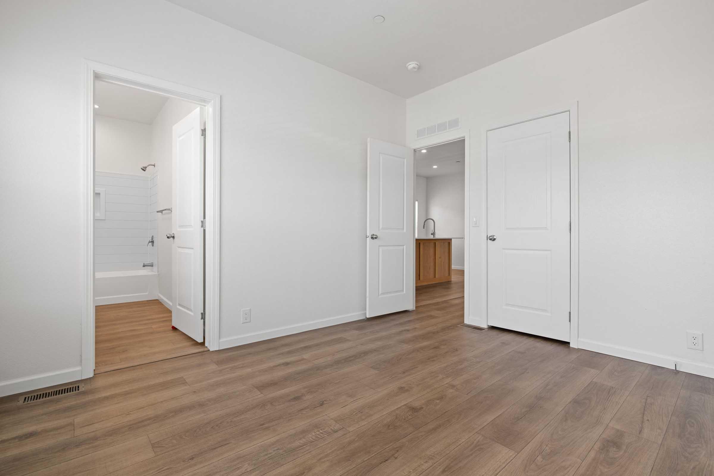 A well-lit room featuring light wood flooring and white walls. Two closed doors are visible, one leading to a bathroom with a tub, and the other possibly to another room. A kitchen area is partially visible in the background. The space has a modern and minimalist design.