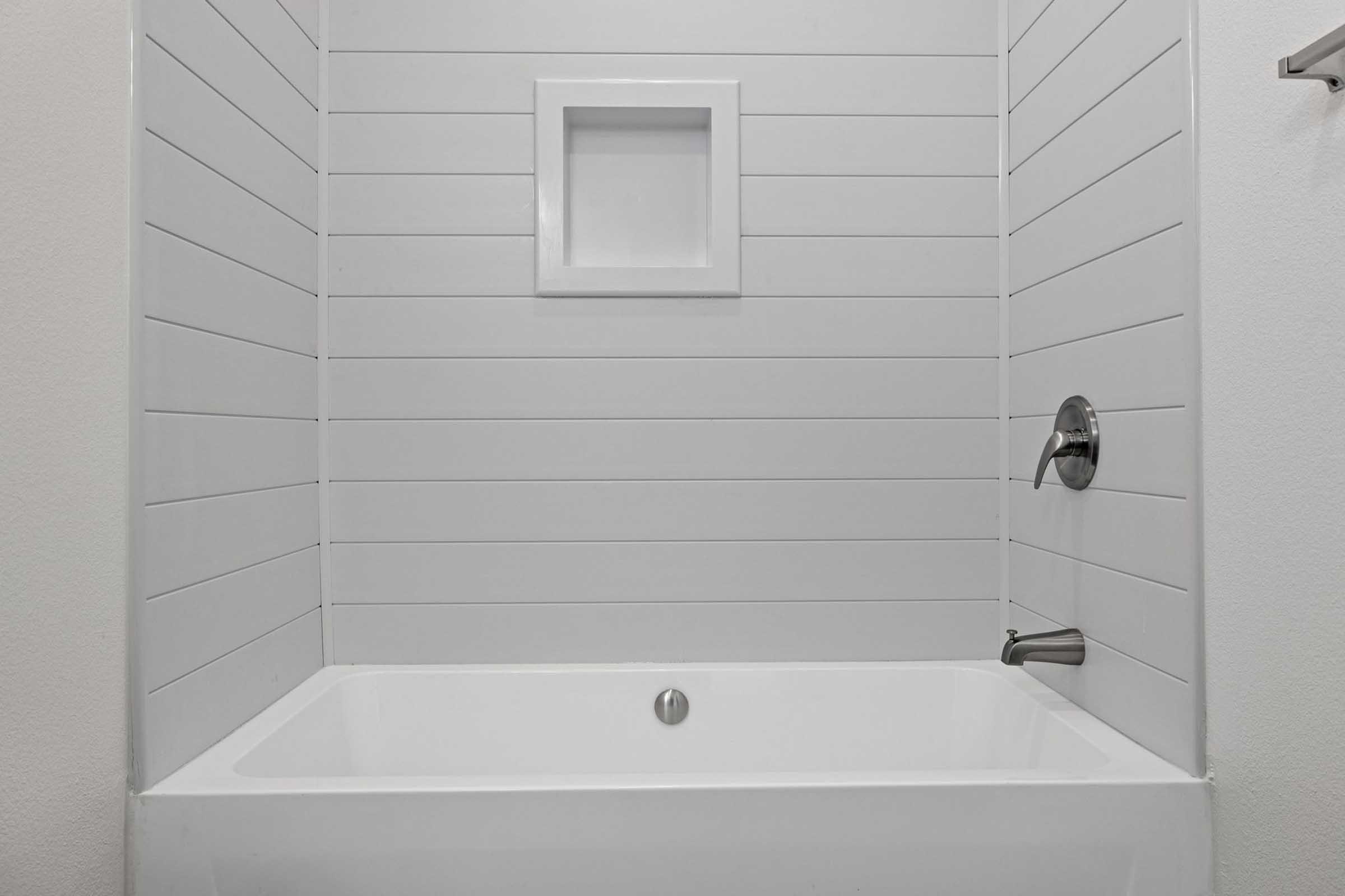 A clean, modern bathtub with white paneling on the walls. There is a built-in shelf for toiletries in the center of the wall. The tub has a sleek faucet and showerhead attached, featuring minimalist design elements in a bright, uncluttered bathroom space.