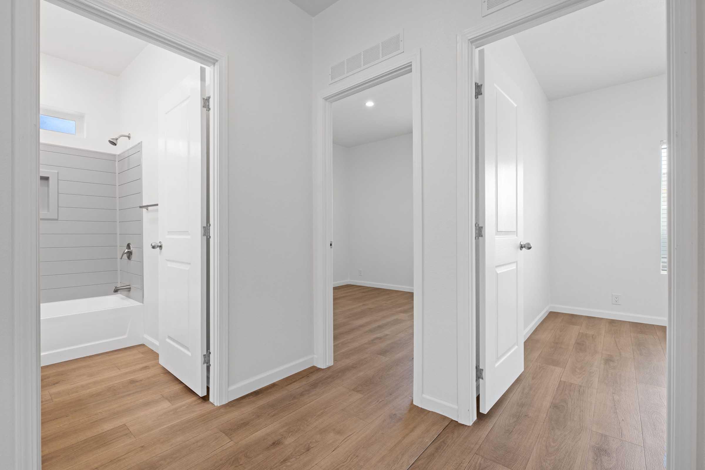 A bright and modern hallway featuring three open doors. Two doors lead to empty rooms, while one door opens to a bathroom with a bathtub visible. The walls are painted white, and the flooring is a warm wood laminate, creating a clean and inviting atmosphere. Natural light illuminates the space.