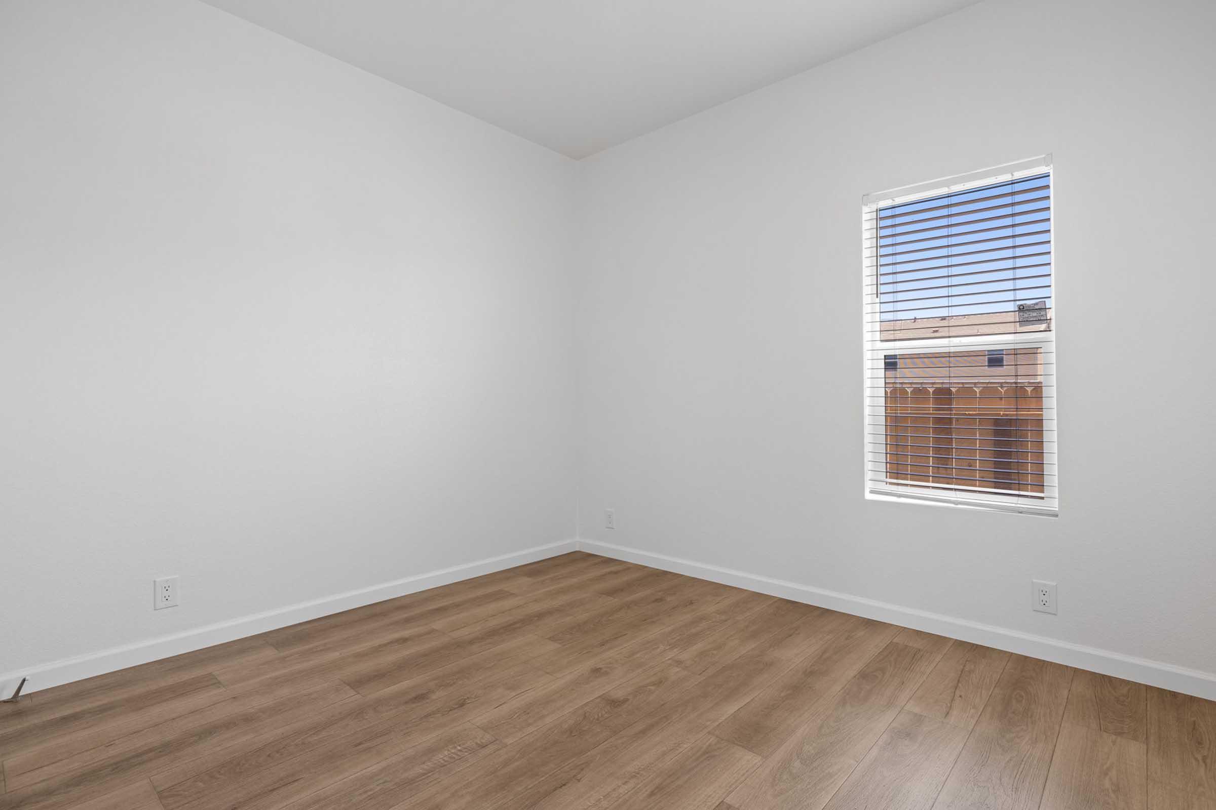 A bright, empty room with white walls and brown laminate flooring. A window with blinds allows natural light to enter, illuminating the space. The room is uncluttered, emphasizing its simplicity and potential for personal decoration.