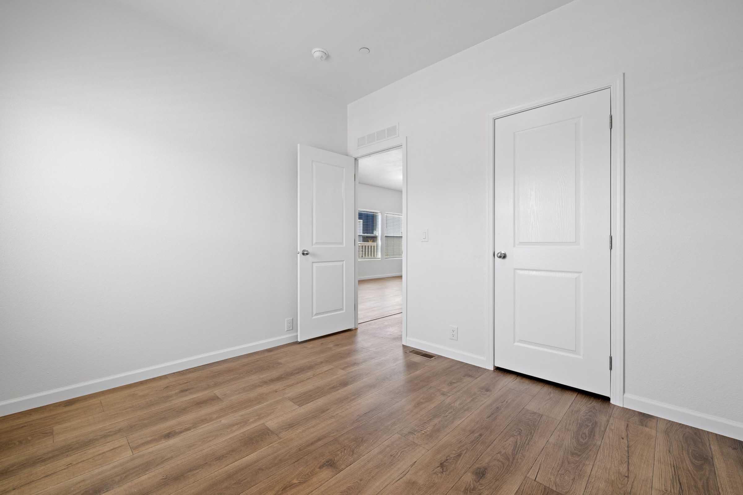 A well-lit, empty room featuring light wooden flooring and white walls. Two doors are visible, one slightly ajar, leading into another area. The room conveys a modern and airy feel, with natural light coming in from a window outside the frame.