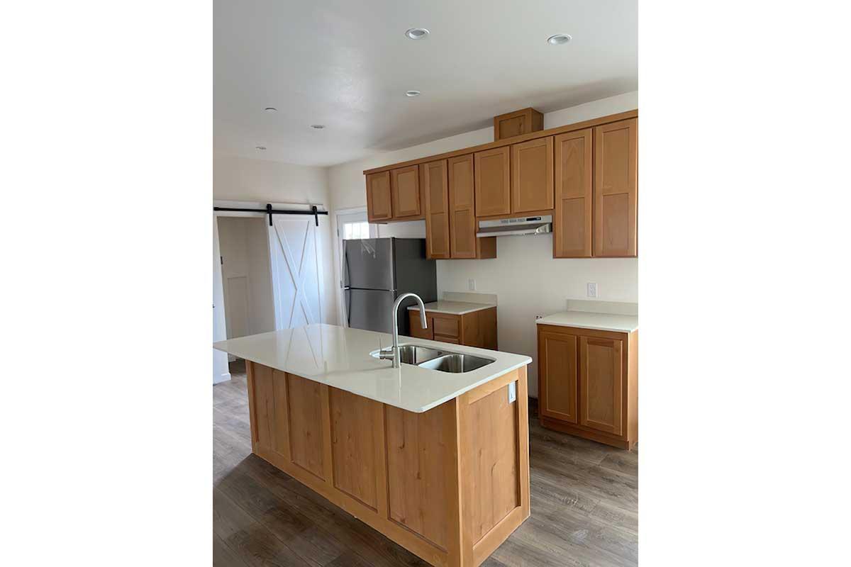 Modern kitchen featuring light brown wooden cabinets, a central island with a white countertop and sink, stainless steel appliances, and a sliding door. The space is well-lit with recessed lighting and has a warm, inviting atmosphere.