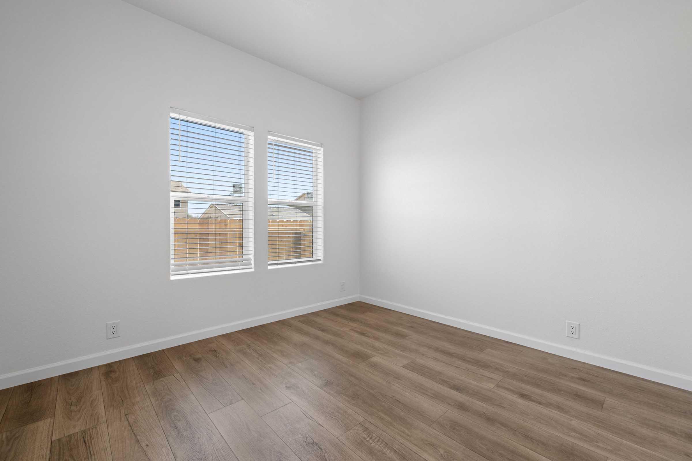 A vacant room with light-colored walls and wooden flooring. It features two windows with blinds that allow natural light to enter, and a view of a fence outside. The space feels clean and bright, ideal for various furnishings or decoration options.