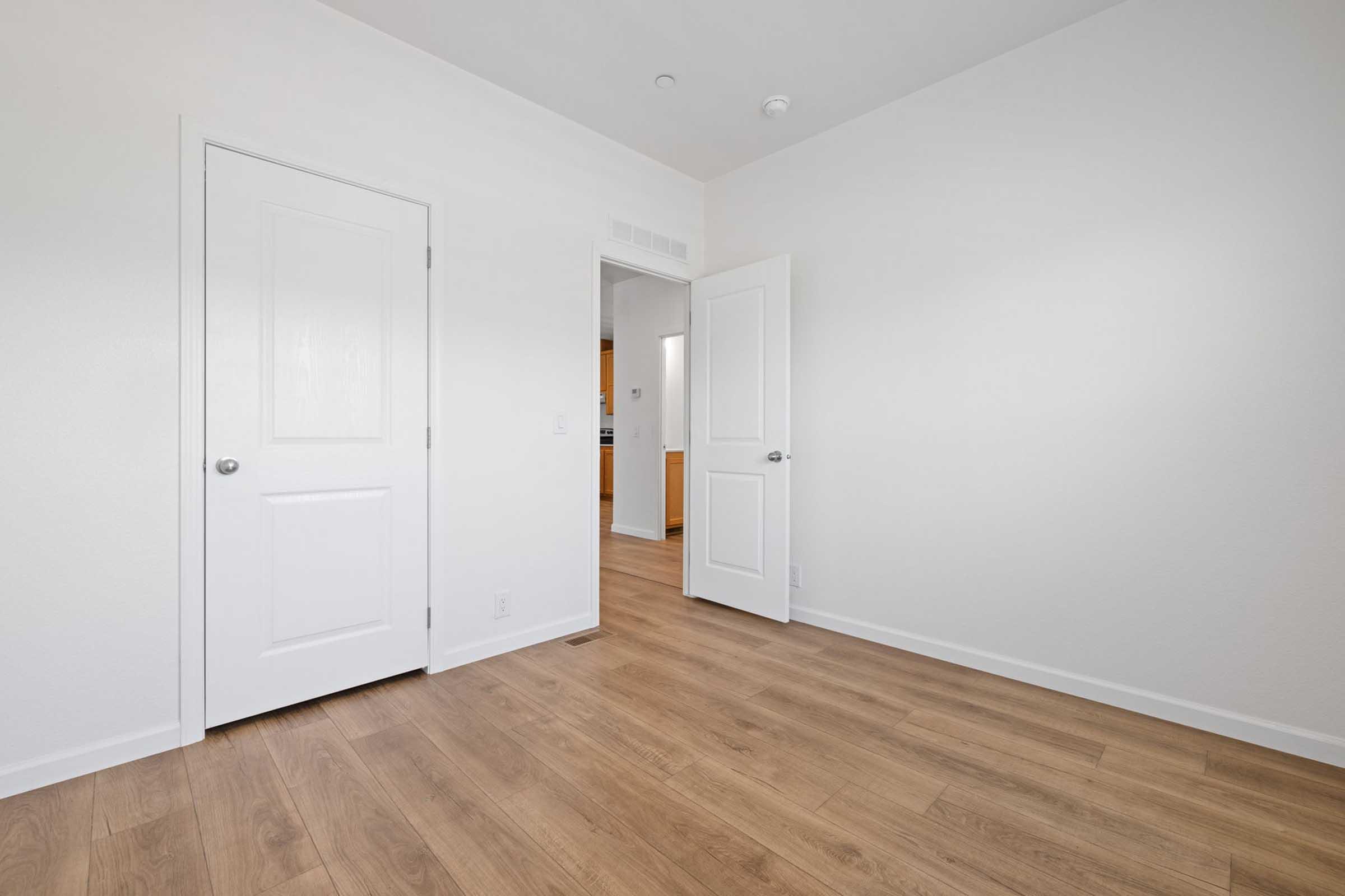 A bright, empty room with light-colored walls and wooden flooring. Two doors are visible: one closed and one slightly ajar, leading to another space. The room has a minimalistic design, emphasizing simplicity and openness.