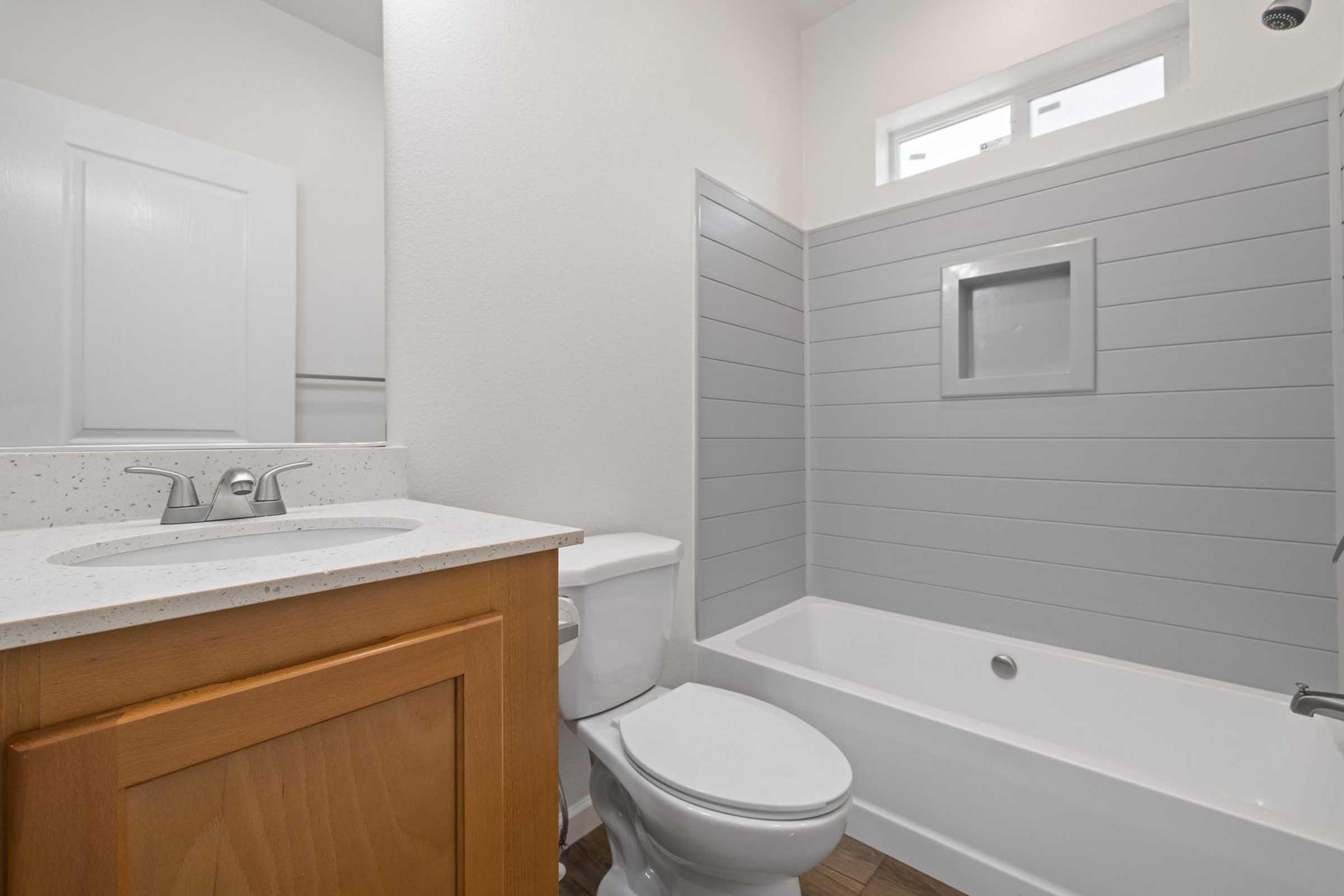 A modern bathroom featuring a bathtub with a shower, light gray shiplap walls, a white sink with a wooden cabinet, and a toilet. The space has a bright and clean design, with natural light coming from a small window above the bathtub.