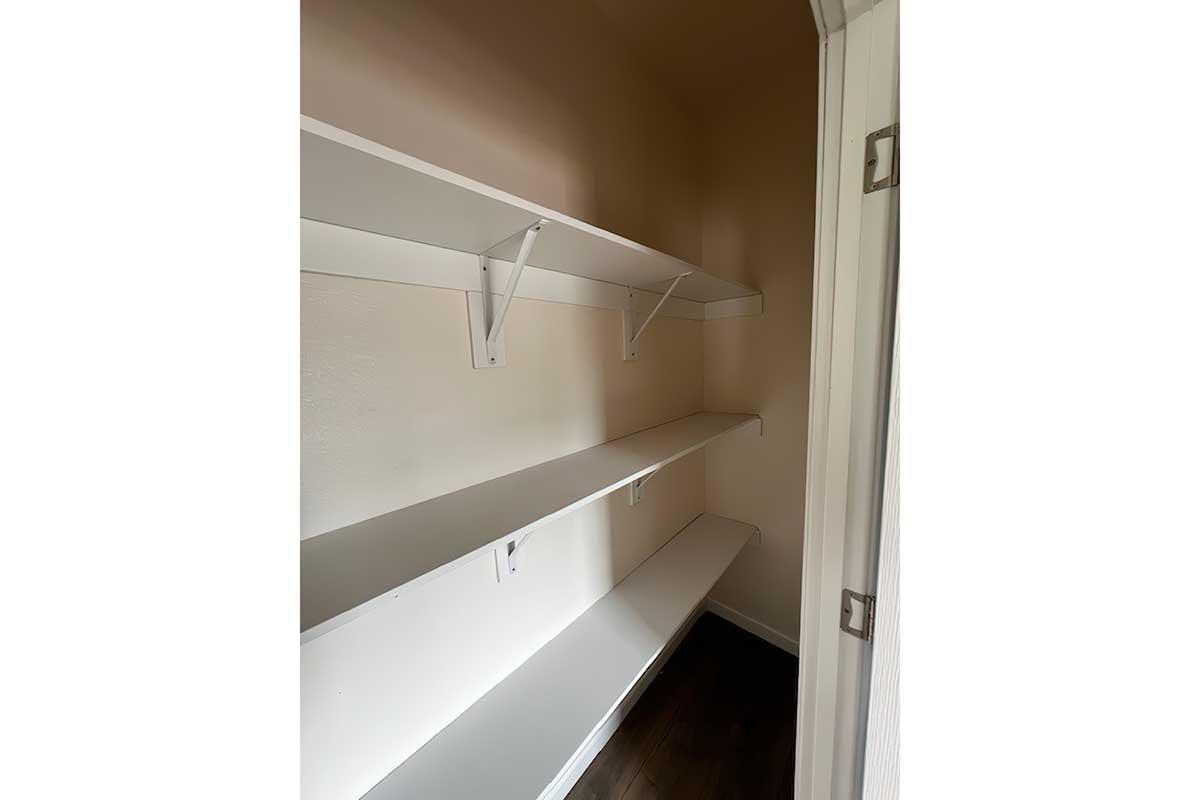 Empty closet with white shelving units mounted on the walls. The shelves are neatly arranged, and the walls are a light, neutral color. The floor is dark, providing contrast to the bright shelves and walls. The space appears clean and organized, suitable for storage or organization.