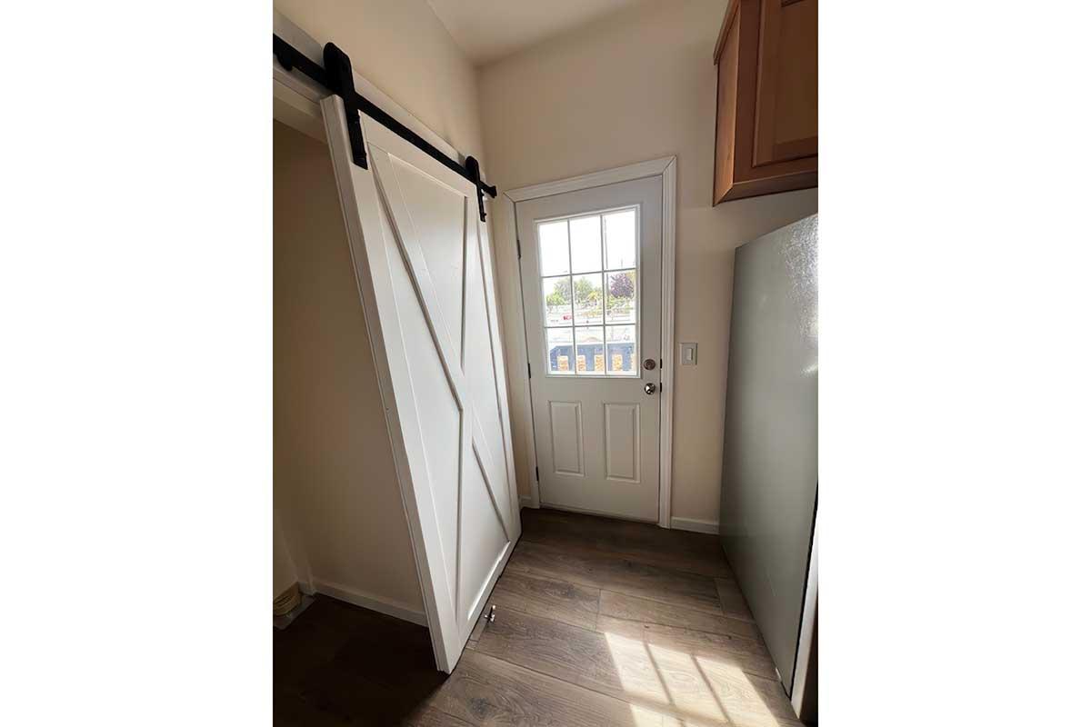 A room featuring a sliding barn door partially open, revealing a door with a window that leads outside. Light-colored walls and wooden flooring are visible, along with cabinetry. The ambiance appears bright and modern.