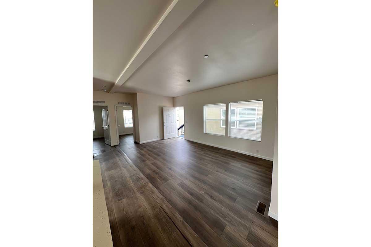 Spacious interior of a modern living room with light-colored walls and hardwood flooring. Large windows provide natural light, and there’s a visible entryway. A kitchen area is partially seen on the left with stainless steel appliances. The overall ambiance is bright and inviting.